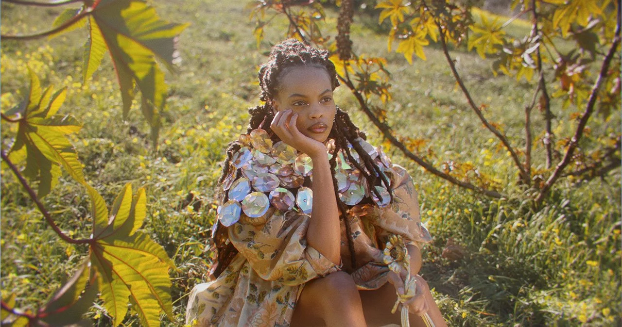 A woman with long dreadlocks sitting among plants and trees, wearing a dress decorated with large reflective disks.