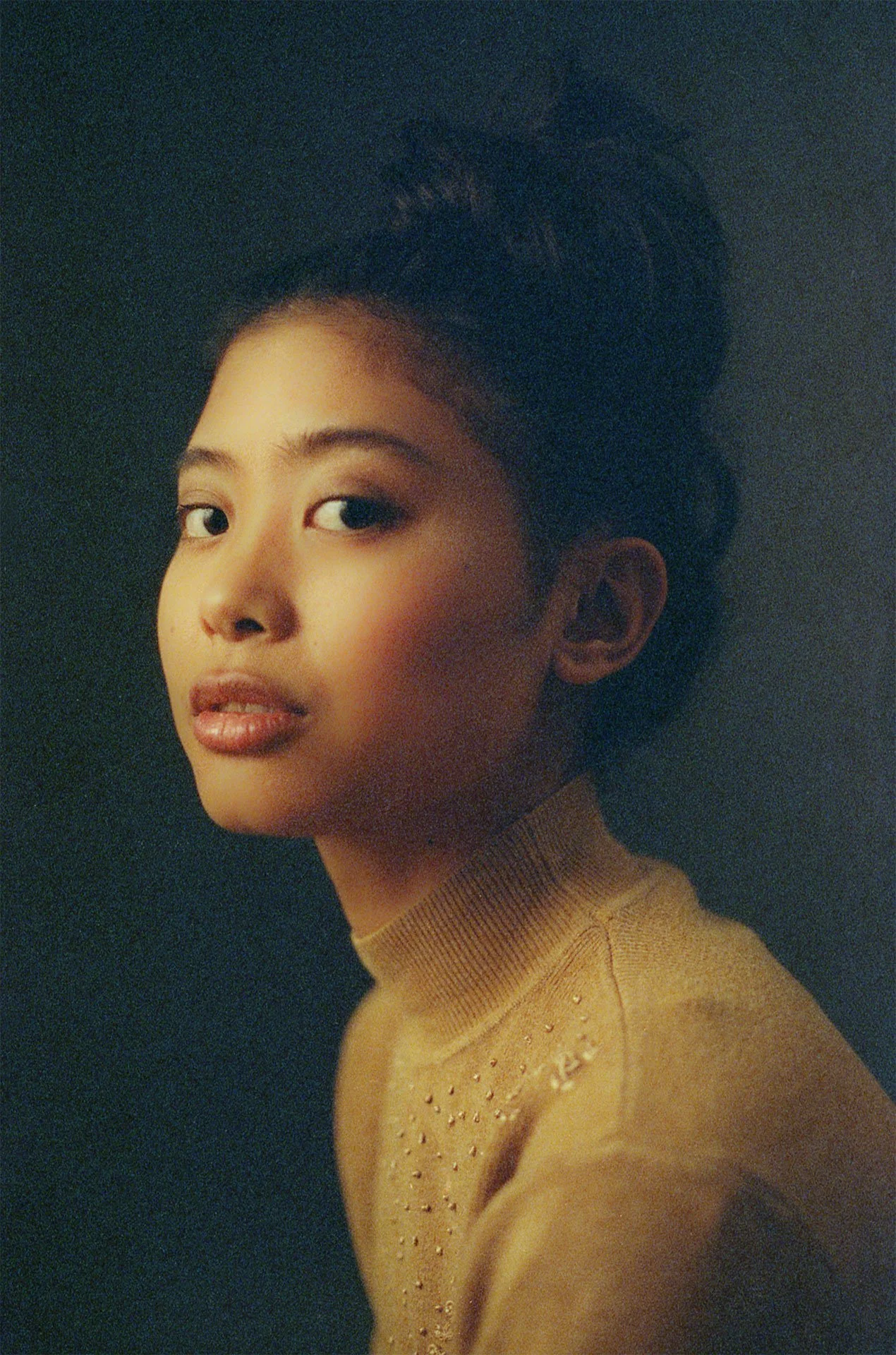 Portrait of a young woman with her hair styled up, wearing a tan turtleneck sweater with small embellishments, against a dark background.