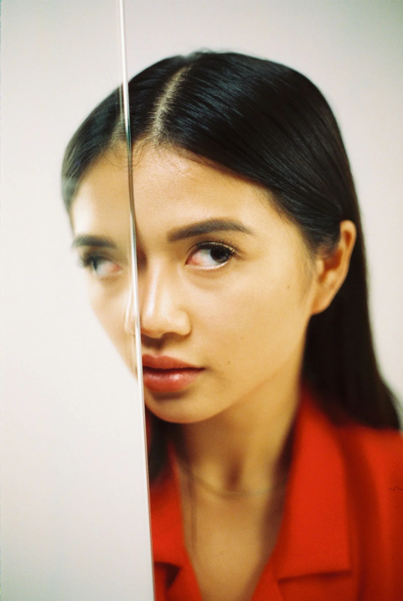 A woman peering around a white wall with a serious expression, wearing a red top and having long, dark, straight hair.