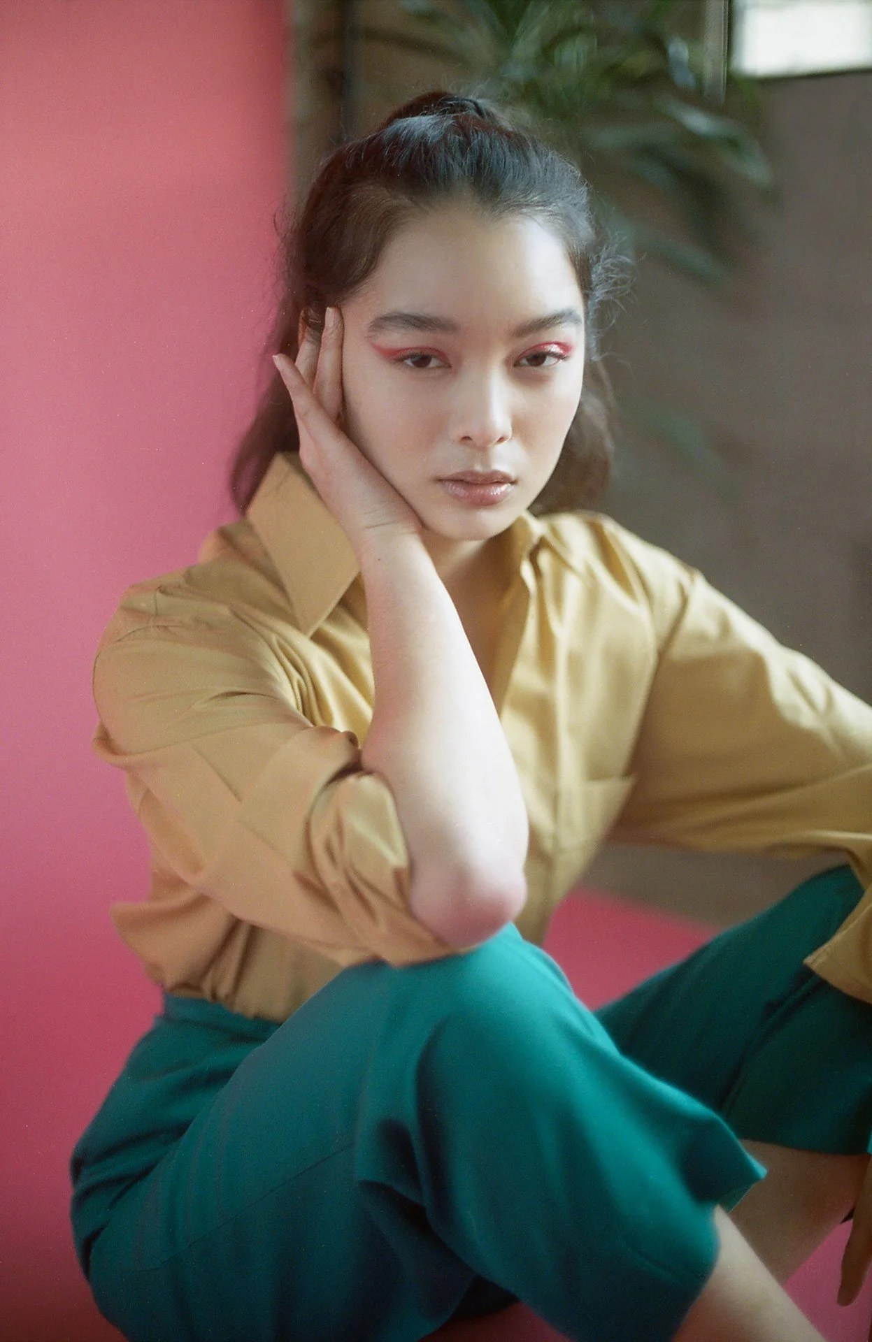 A young woman sitting with her head resting on her hand, wearing a beige shirt and teal pants, with a pink wall and some green plants in the background.
