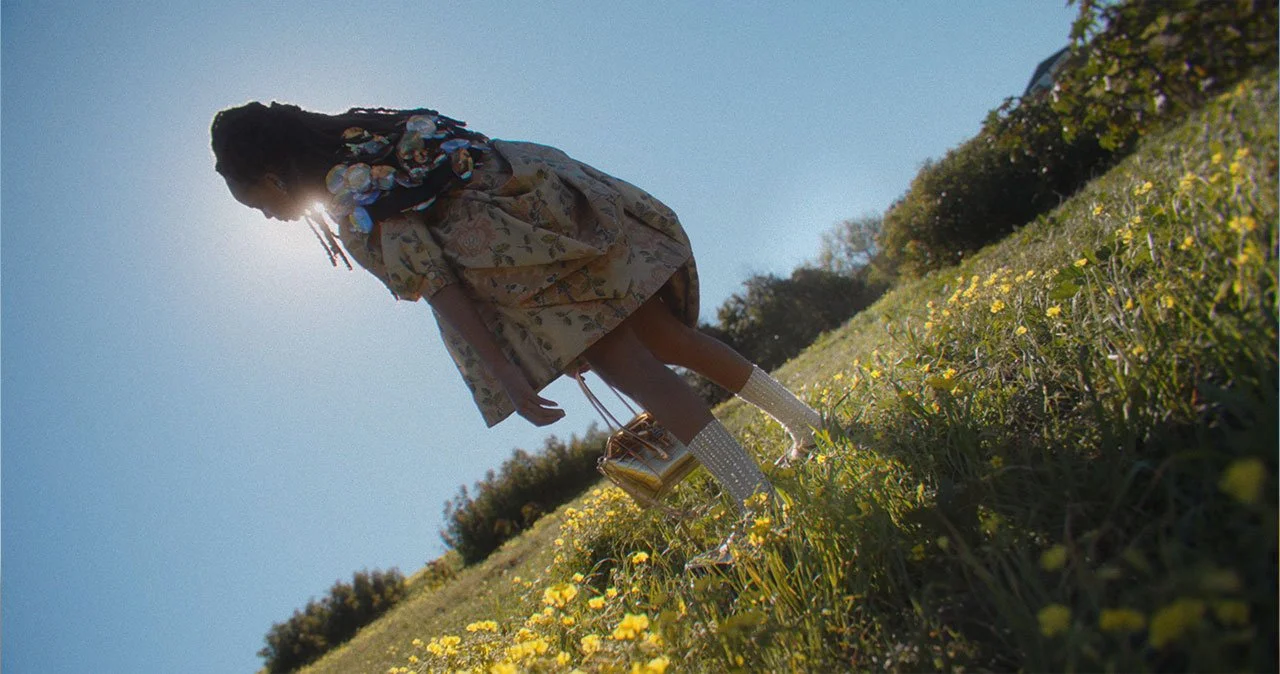 A girl with dreadlocks wearing a floral dress and sparkly knee-high socks standing in a field of yellow flowers with a clear blue sky in the background.