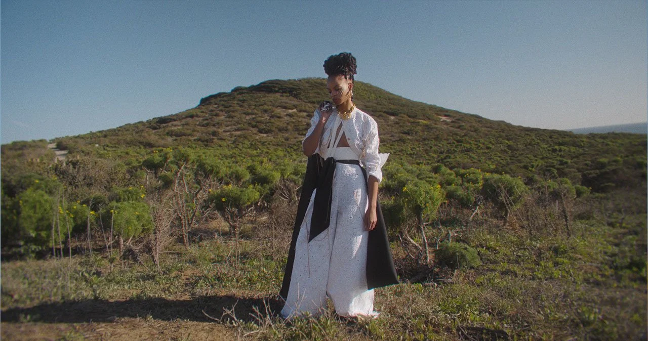 A woman standing outdoors in a hilly, shrub-covered landscape under a clear sky, wearing a white and black outfit with jewelry and an updo hairstyle.