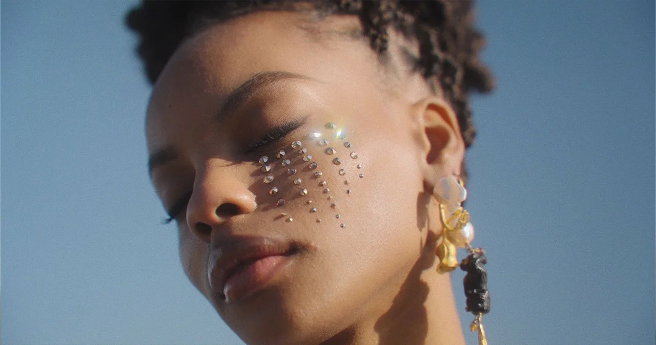 Close-up of a woman with decorated face, wearing large earrings, with her eyes closed against a blue sky.