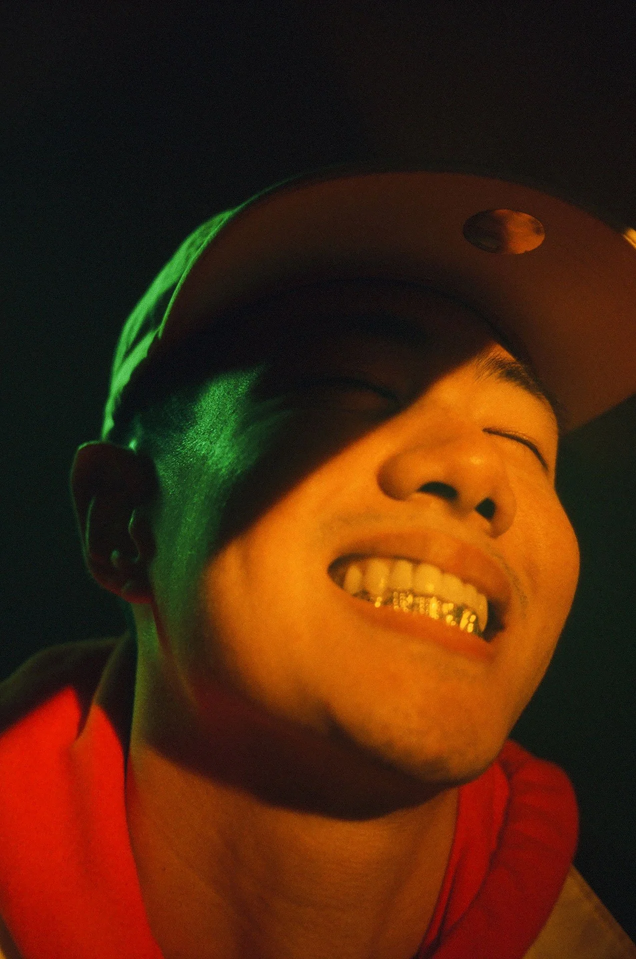 Close-up of a smiling young man wearing a baseball cap, showing his teeth with grillz, against a dark background.