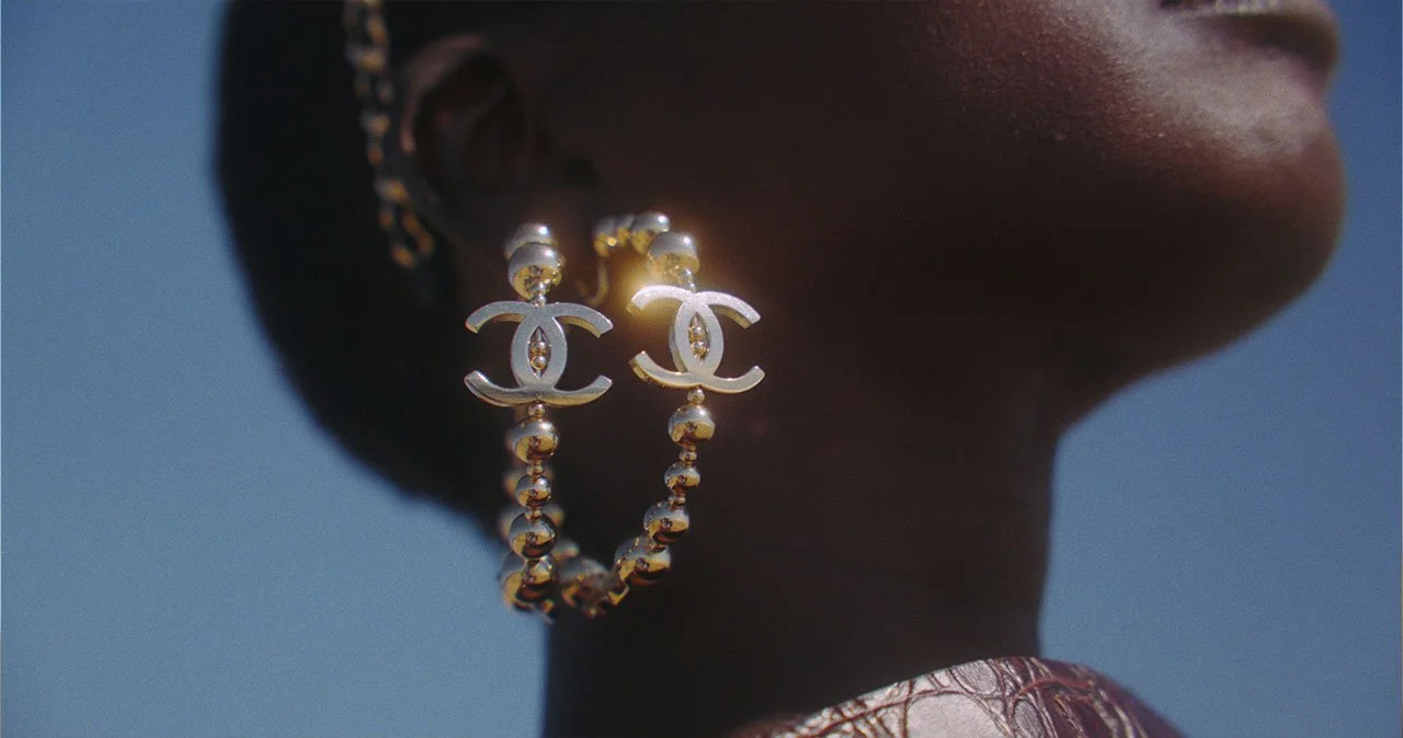 Close-up of a person's face showing only the lower half, wearing gold earrings with the Chanel logo and a beaded necklace, against a blue background.