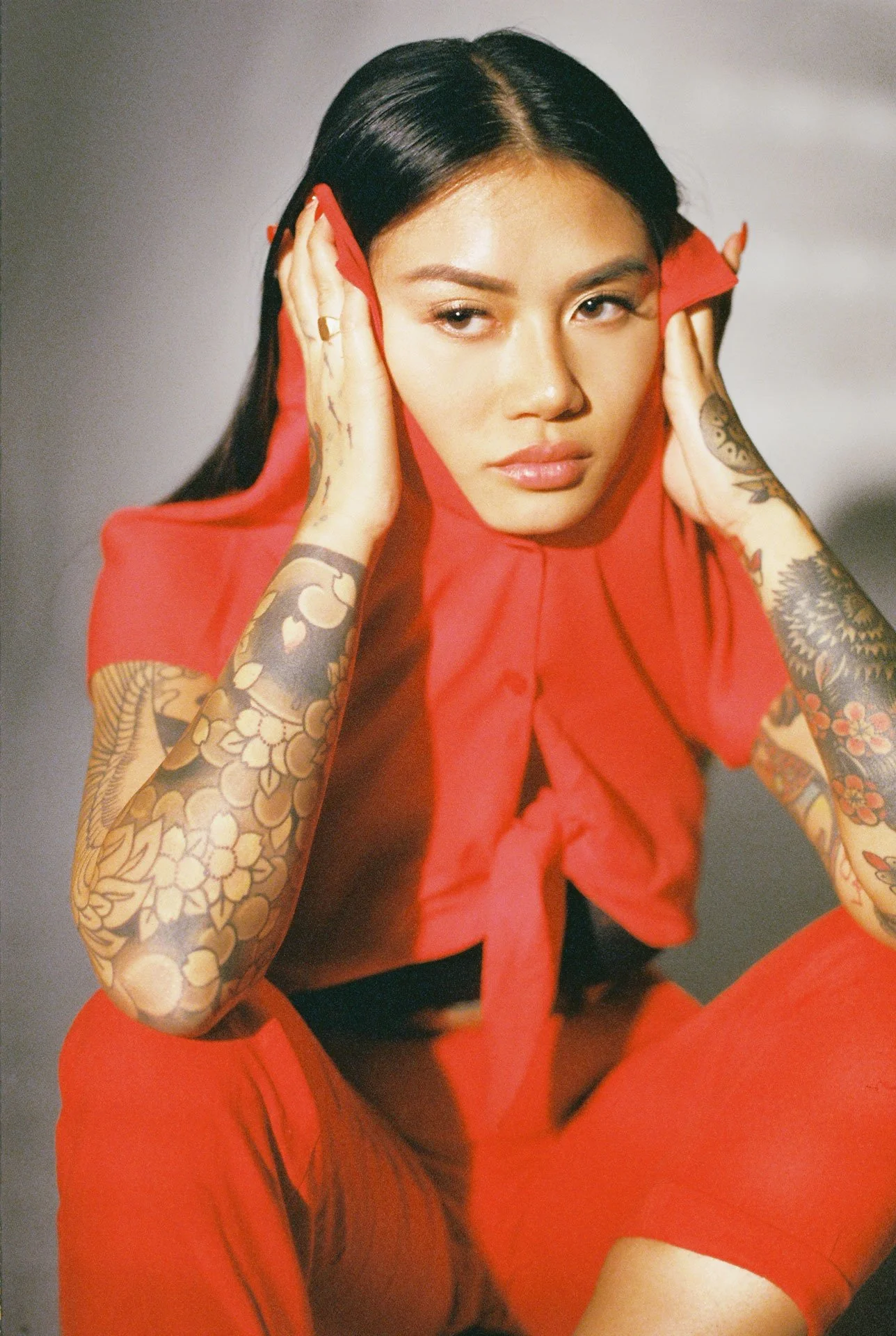 A woman with dark hair and tattoos on her arms, wearing a red outfit, is holding her head with both hands and looking at the camera with a serious expression.