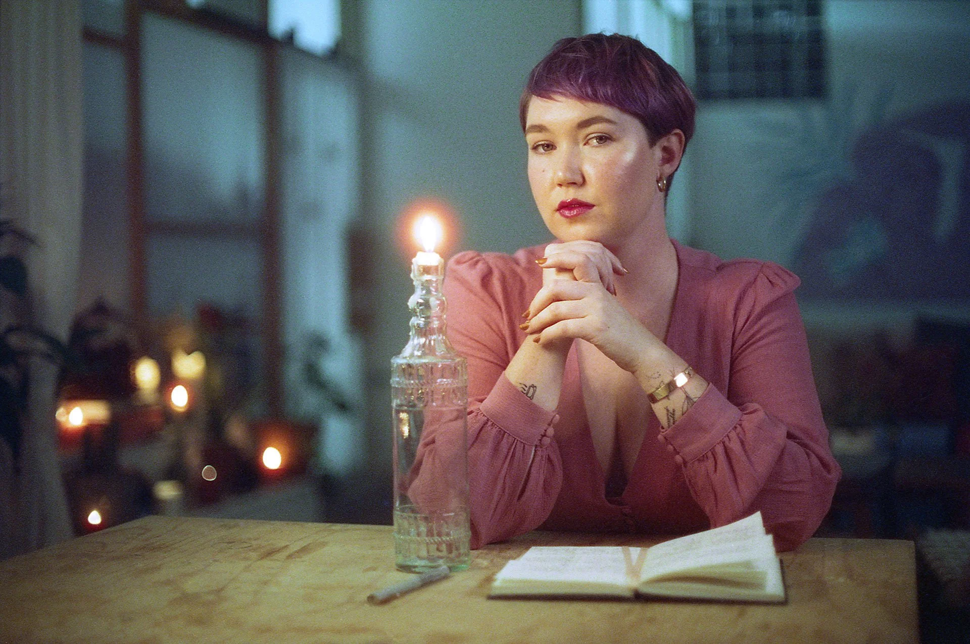 A woman with short purple hair, wearing a pink long-sleeve top, sits at a wooden table with a green backdrop. She is resting her chin on her clasped hands and looks at the camera. There is a lit candle in a glass bottle, an open notebook, and a pen on the table.