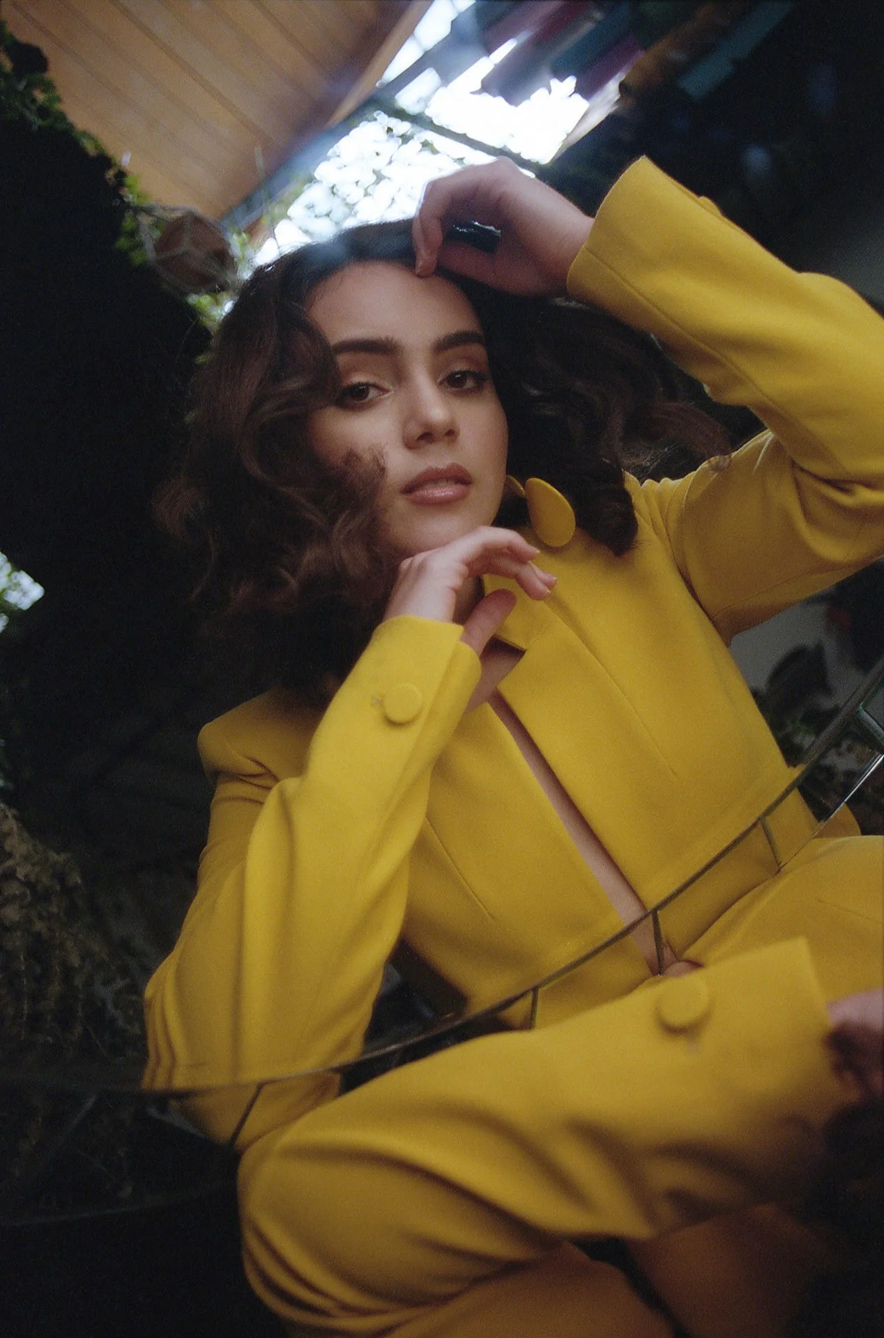 A young woman with curly hair wearing a yellow blazer with large buttons, looking into a mirror while posing with her hand near her face. The background suggests an indoor setting with plants and natural light.
