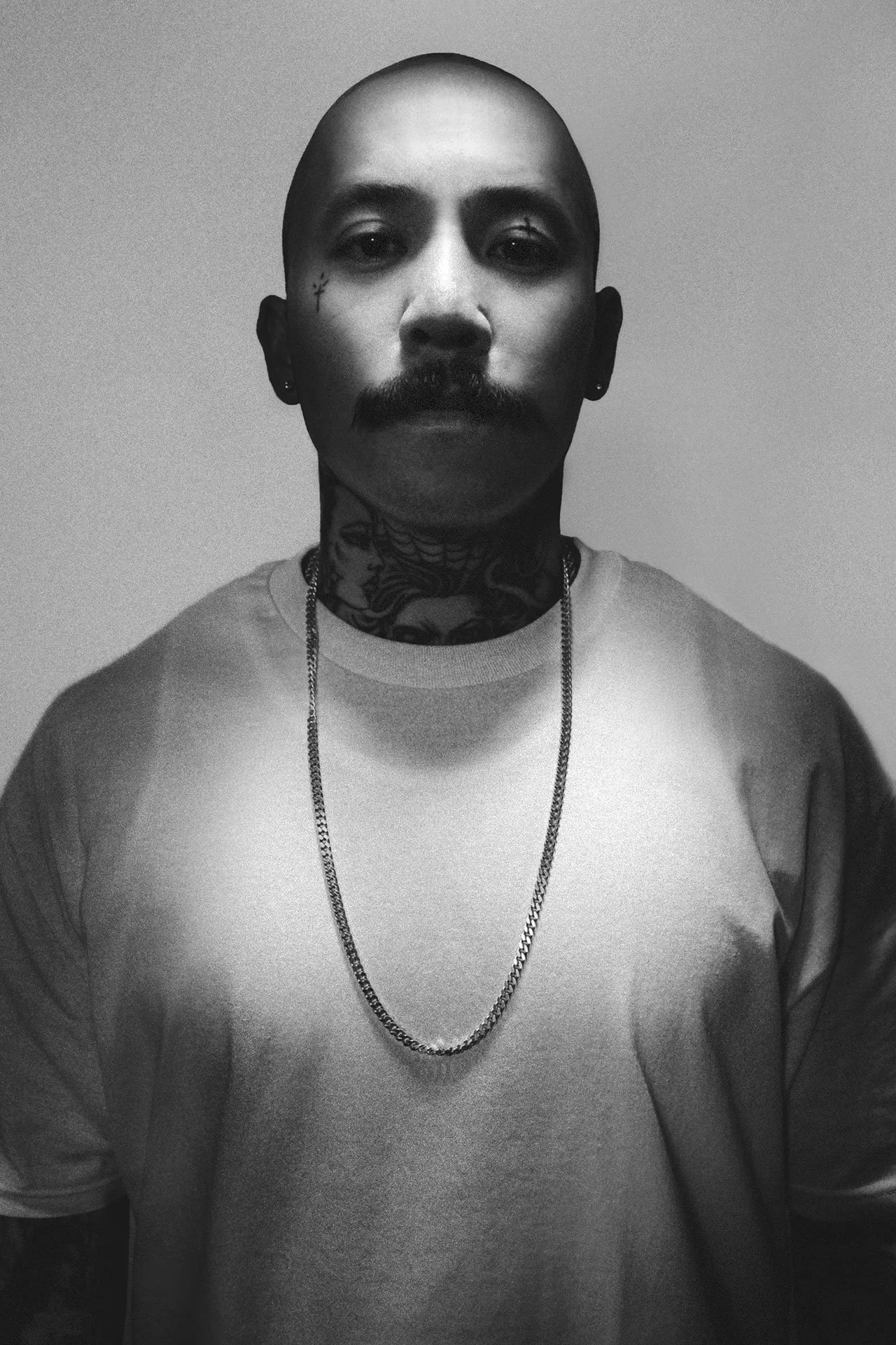 A black and white portrait of a man with tattoos, a mustache, and a shaved head, wearing a plain T-shirt and a chain necklace, standing against a plain background.