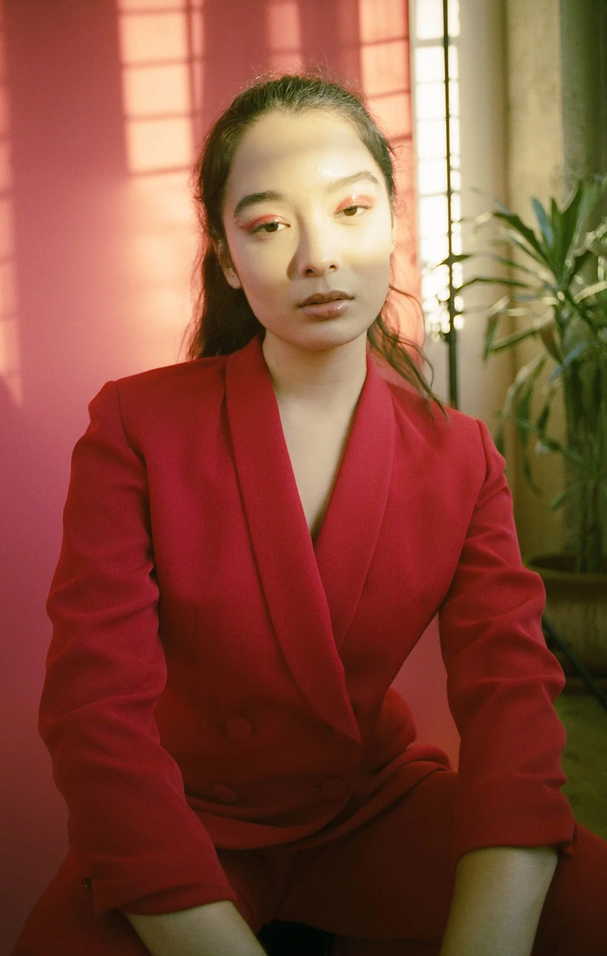 A woman with dark hair tied back, wearing a red blazer, sitting in front of a pink wall with sunlight casting shadows and a window with blinds behind her.