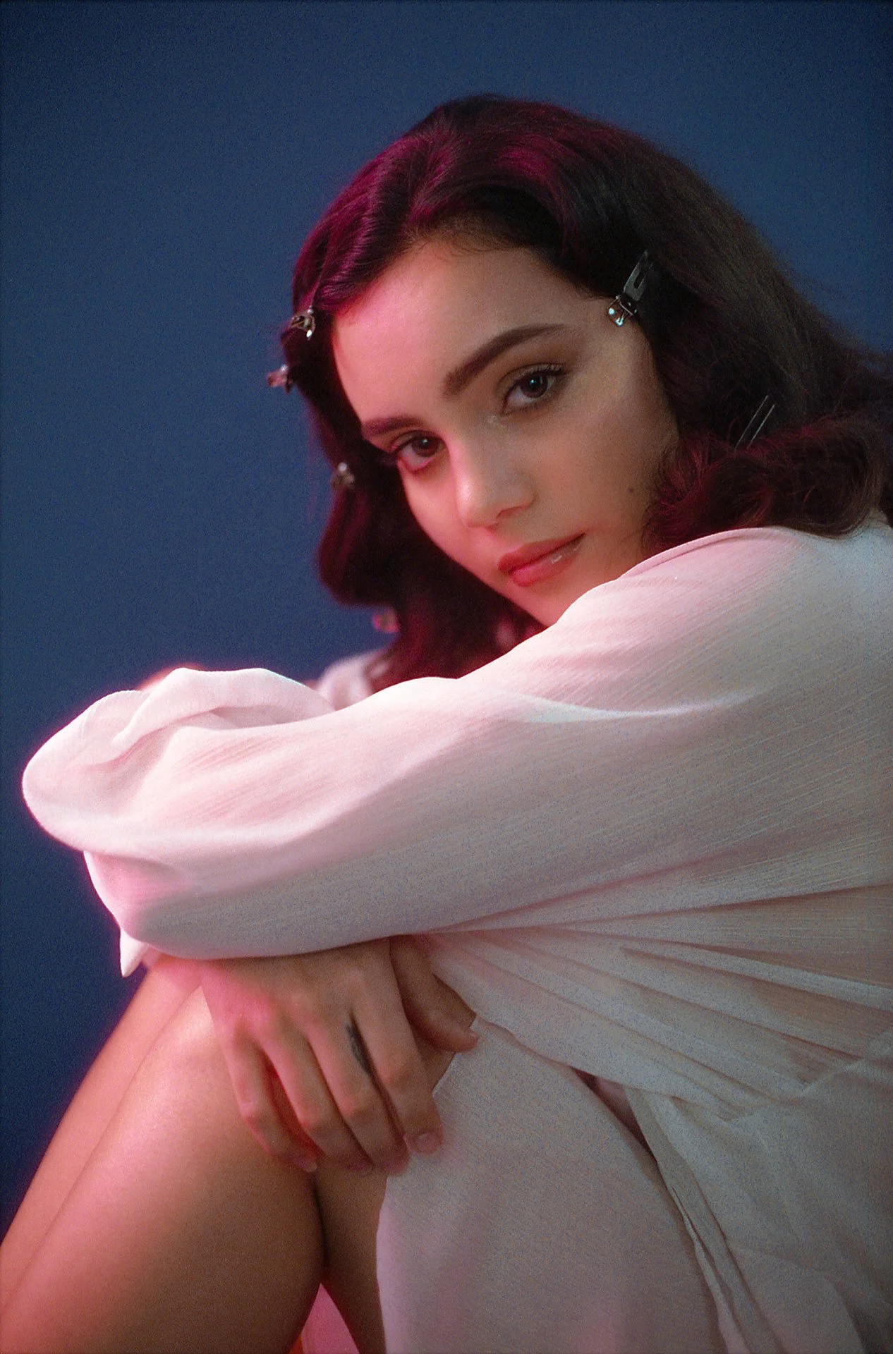 A young woman with dark, wavy hair accented with hair clips, wearing a white long-sleeve top, looking at the camera with a gentle expression, against a plain dark background.