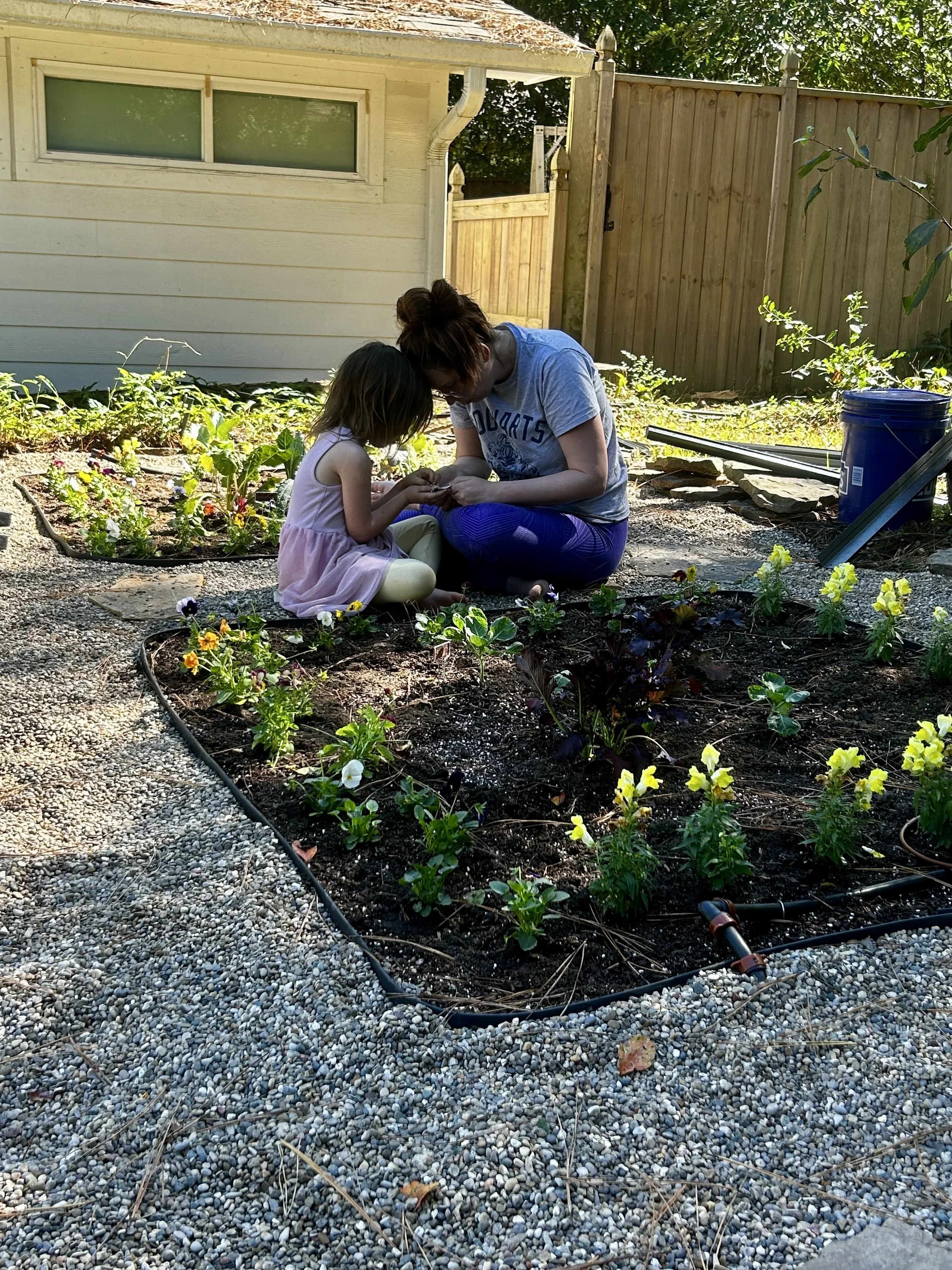 A backyard patio with a gravel ground, surrounded by greenery, featuring three Adirondack chairs around a fire pit, with a potted succulent in the foreground and a wooden fence in the background.