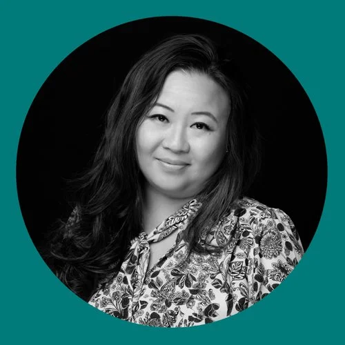 A black and white portrait photograph of a woman with long dark hair, smiling, wearing a patterned blouse, set against a circular backdrop with a teal green background.