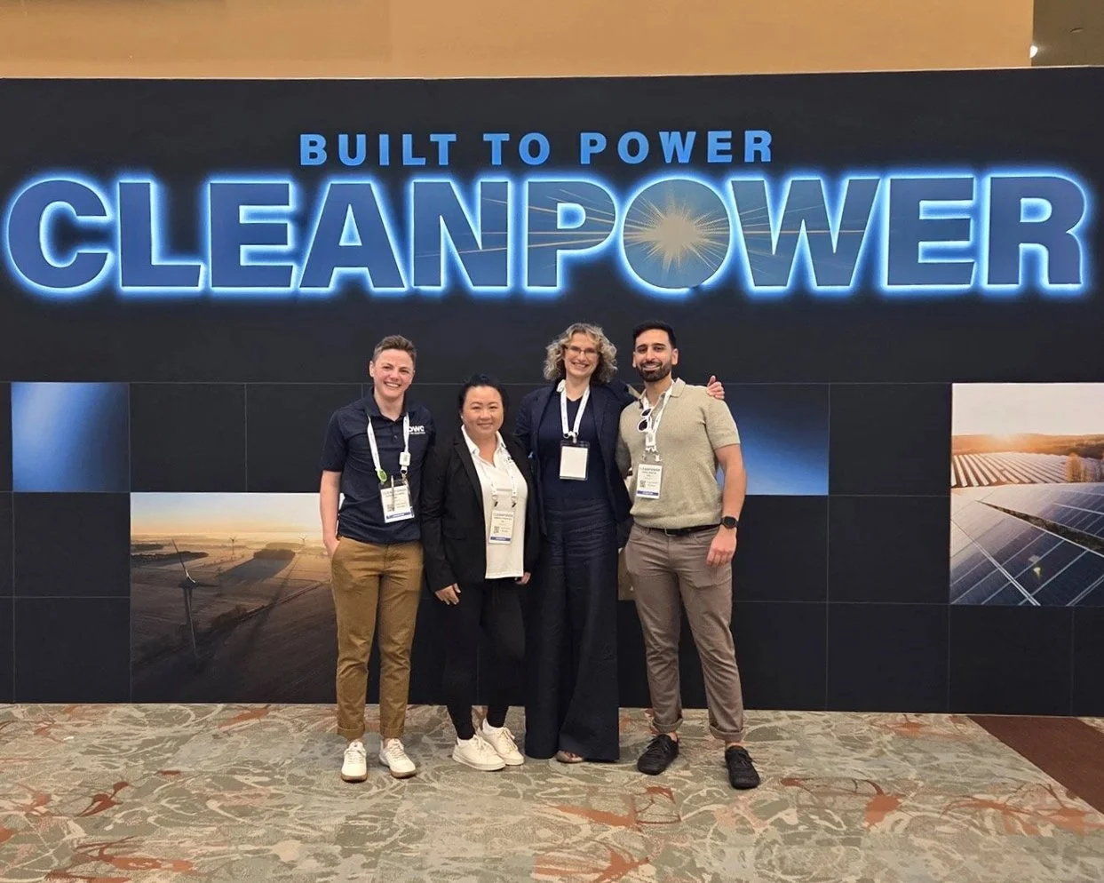 Group of four people standing in front of a large sign that says 'BUILT TO POWER CLEANPOWER' at an event, with two women and one man smiling, wearing conference badges and casual attire.