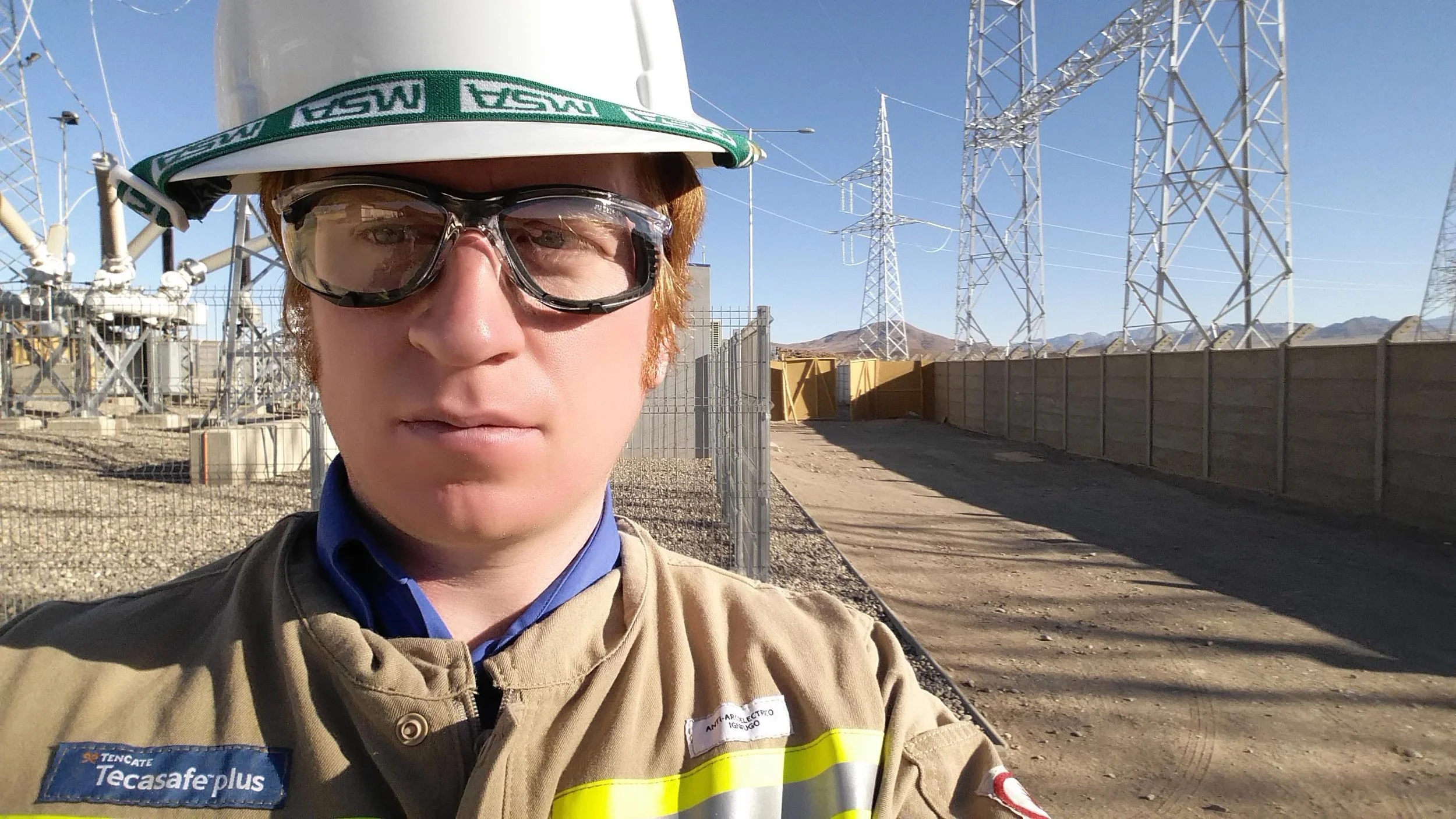 A person wearing safety goggles, a hard hat with a green strap, and a work uniform taking a selfie in an industrial or electrical substation area with power lines and towers in the background.