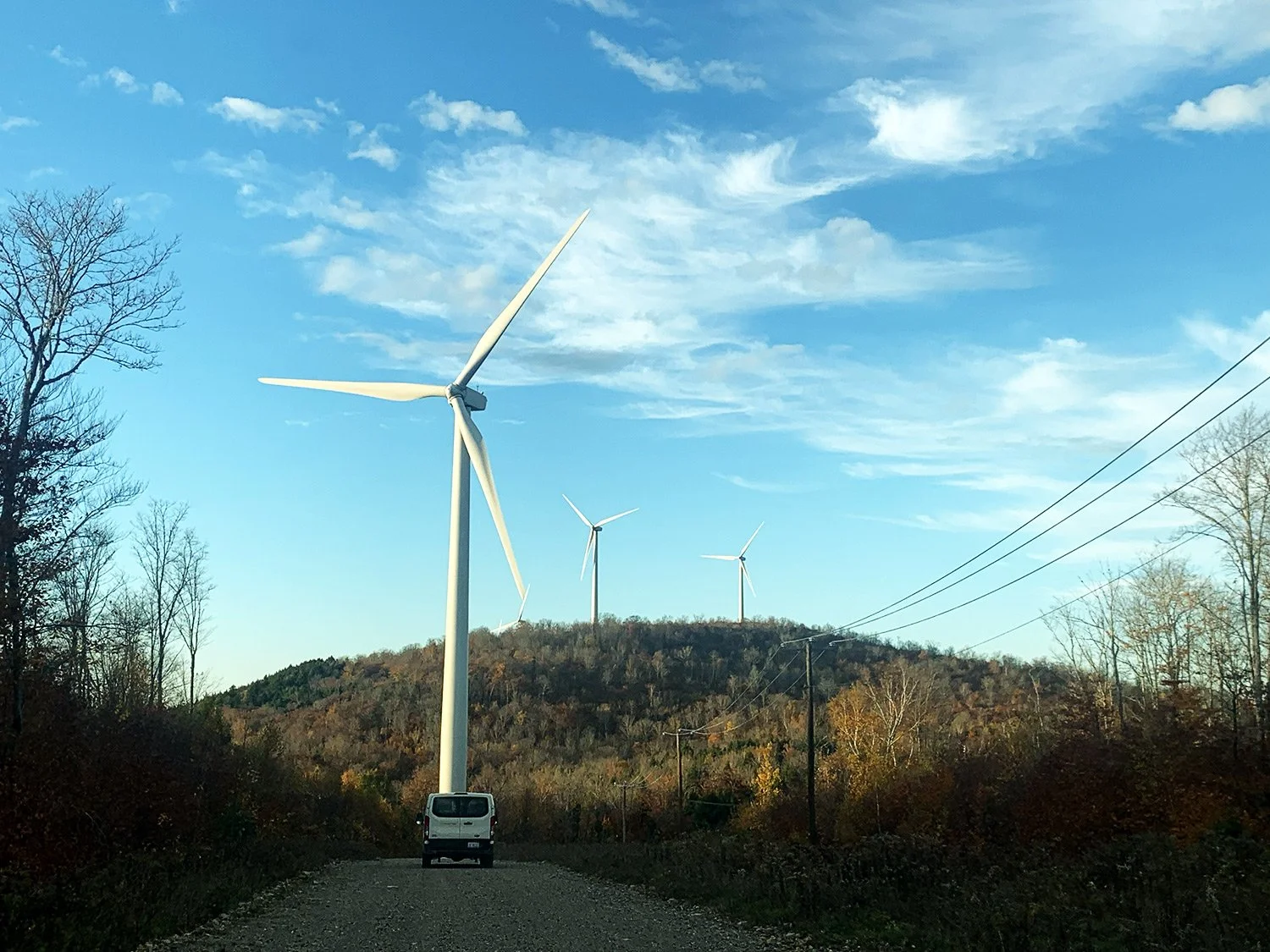 A gravel road leading towards wind turbines on a hill with trees in fall colors, power lines on the right, and a clear blue sky with clouds.