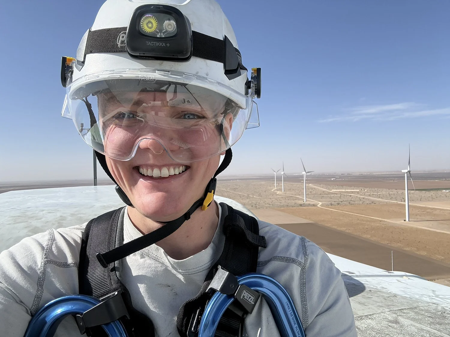 A person smiling at the camera wearing safety gear, including a helmet with a headlamp and goggles, standing on top of a structure in a wind farm with wind turbines in the background in a flat, arid landscape under a clear blue sky.