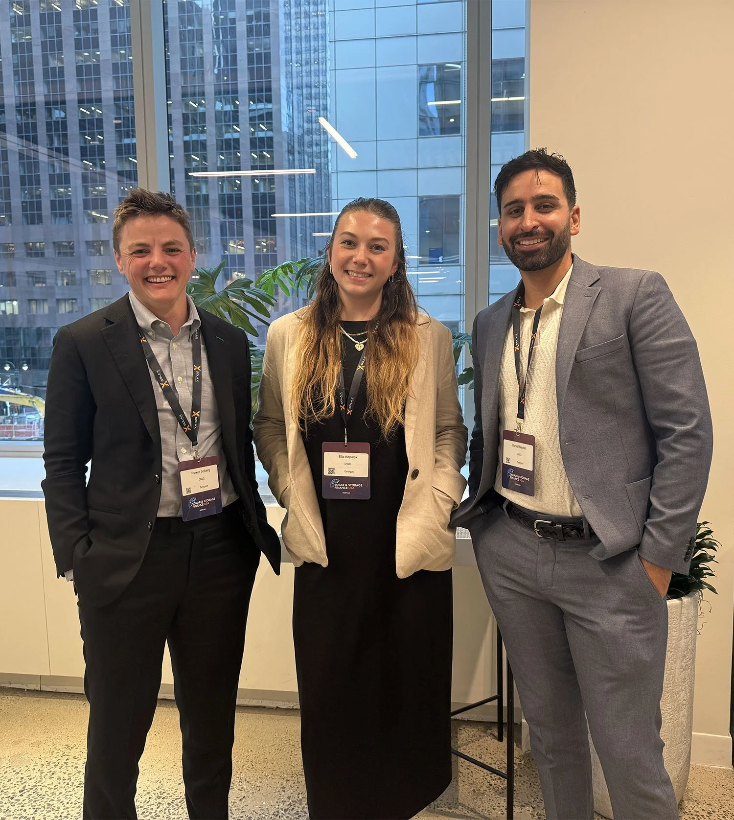 Three smiling professionals standing together in an office with large windows and a cityscape view, wearing business attire and conference badges.