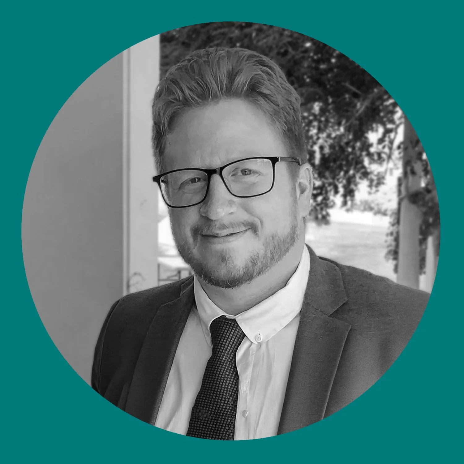Black and white photo of a man wearing glasses, a suit, and a tie, smiling, outdoors with trees in the background, framed within a circular teal design.