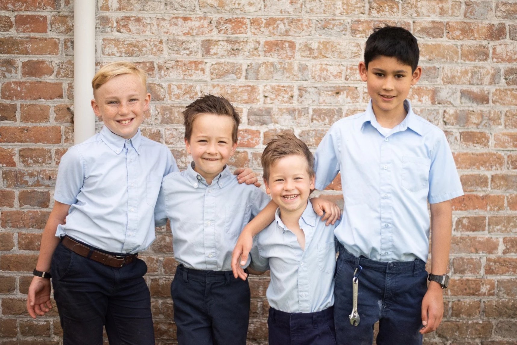 Four boys in light blue button-up shirts and dark pants standing together against a brick wall, smiling for a group photo.