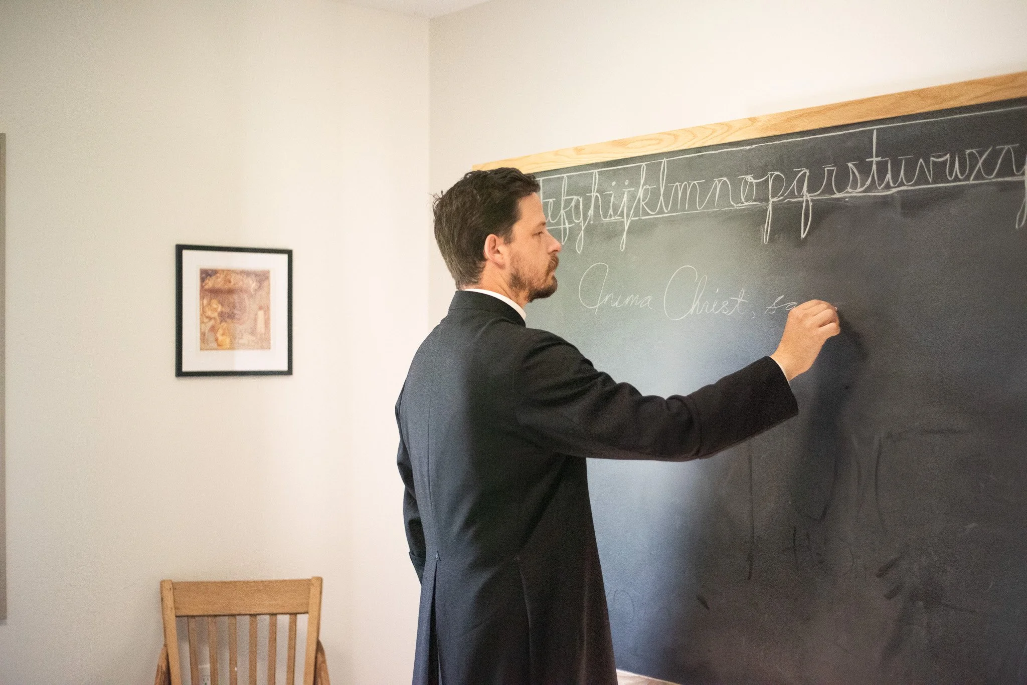 A priest in a cassock writing on a black chalkboard in a classroom.