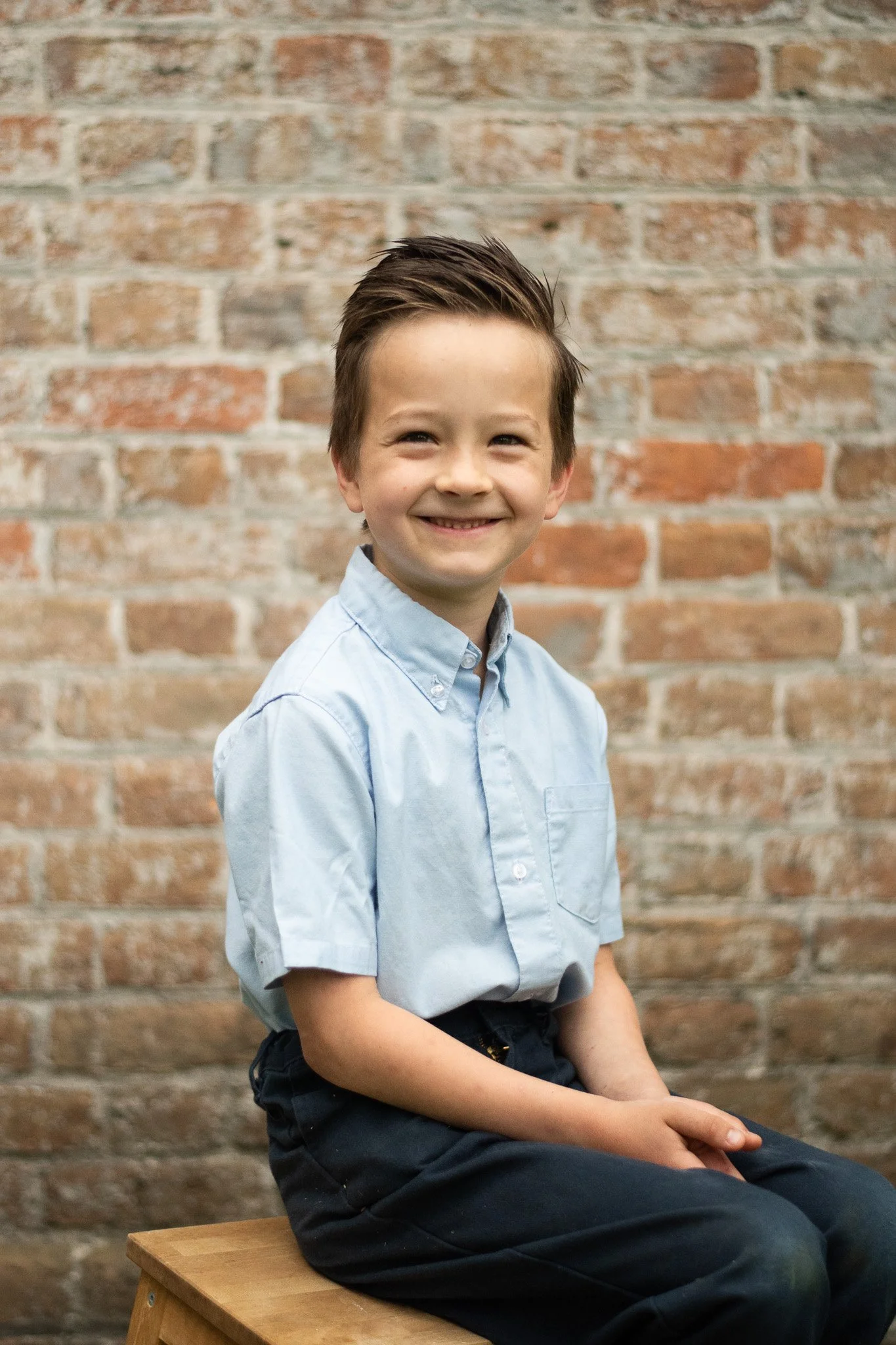 A young boy with short brown hair smiling, wearing a light blue button-up shirt and black pants, sitting on a wooden stool against a brick wall background.