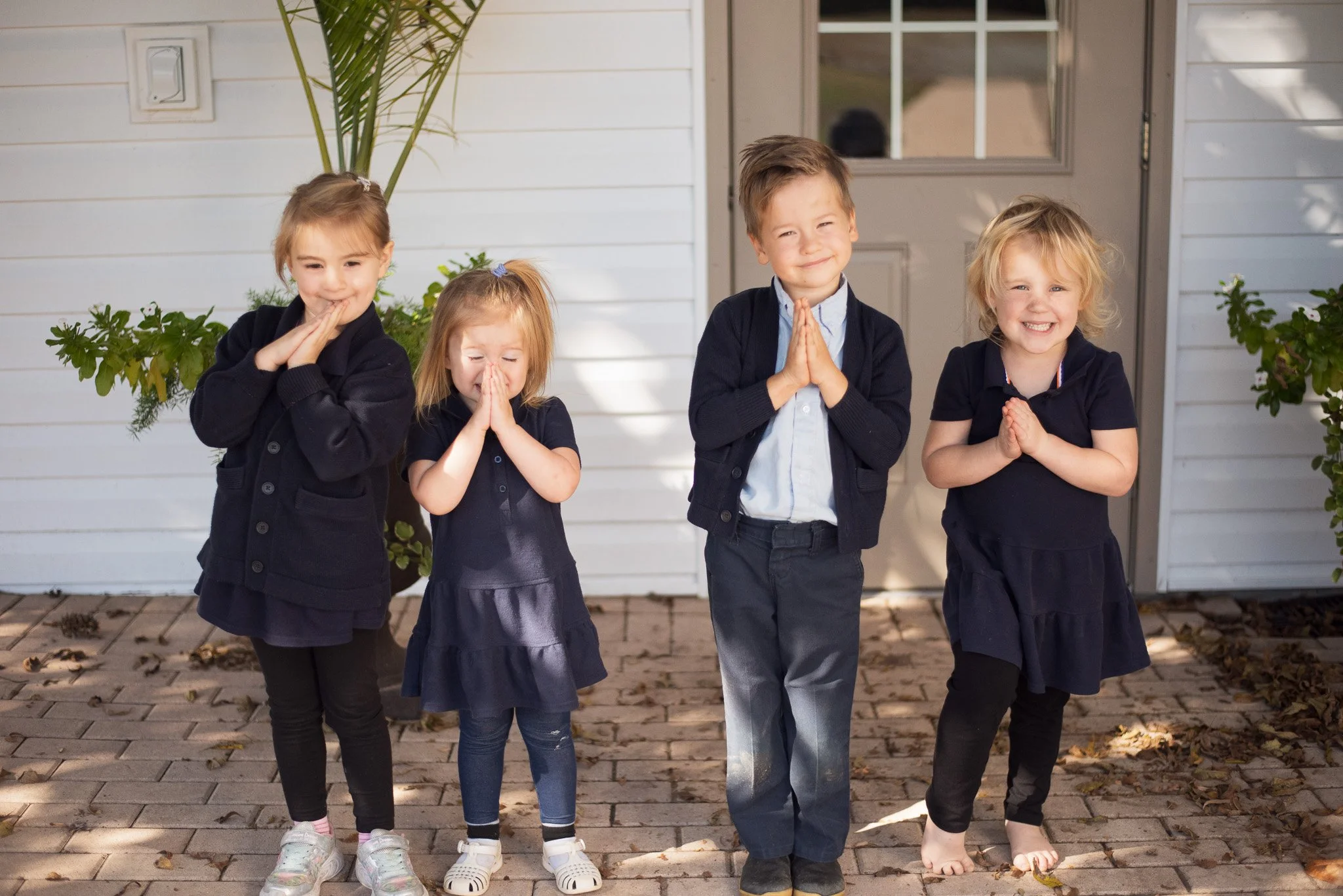Four children standing outdoors on a paved area with faint autumn leaves, in front of a white house with a window and door, holding their hands together in prayer or greeting, smiling at the camera.