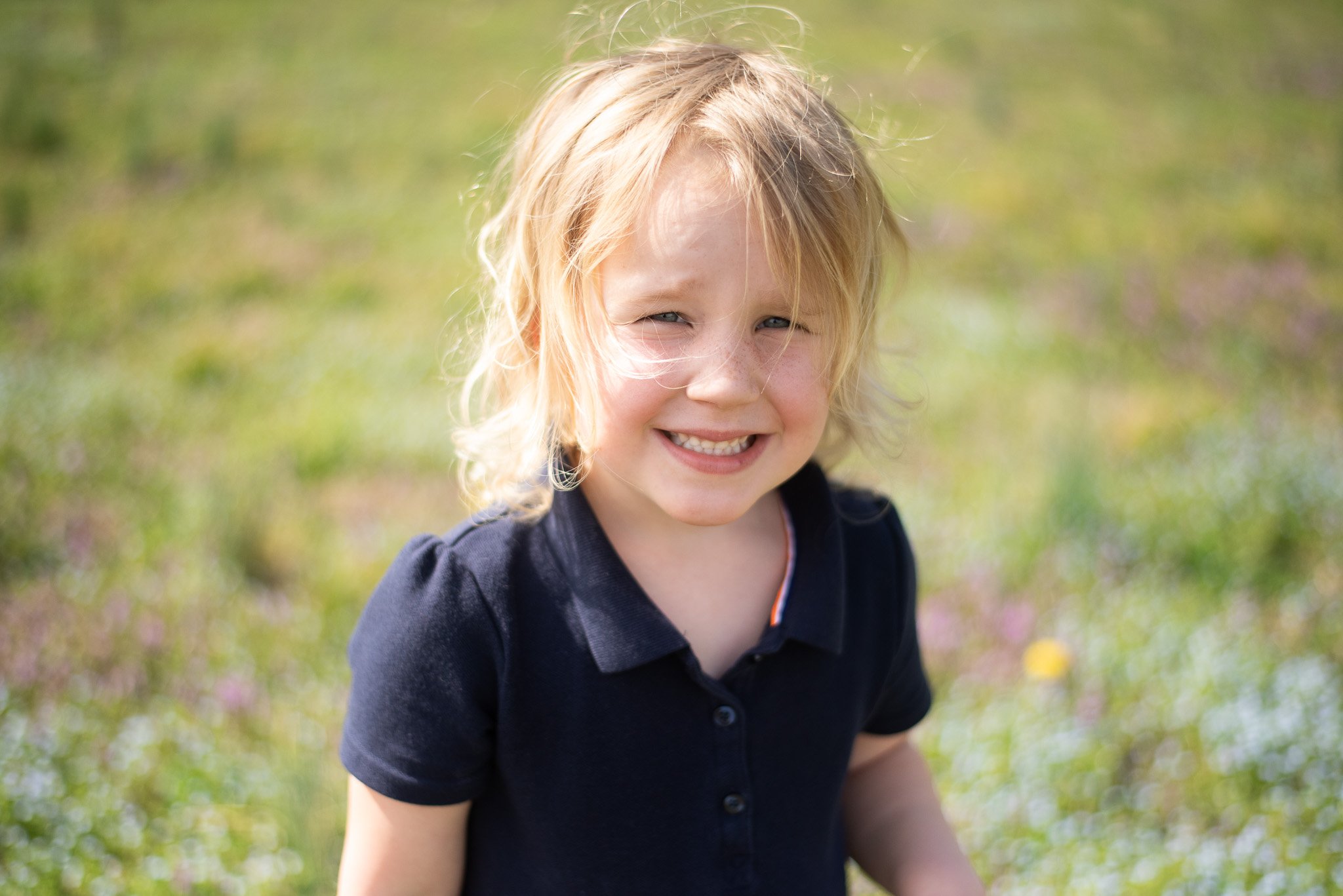 A young girl with blonde hair, smiling outdoors in a grassy area with flowers.