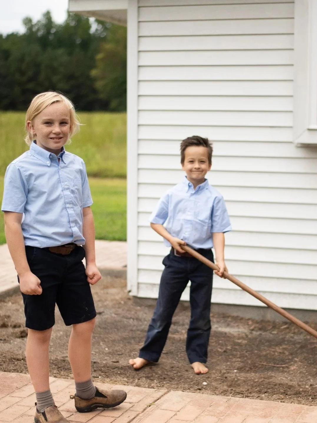 Two young boys are working outside, one holding a rake and the other standing nearby, in front of a white house with green trees in the background.