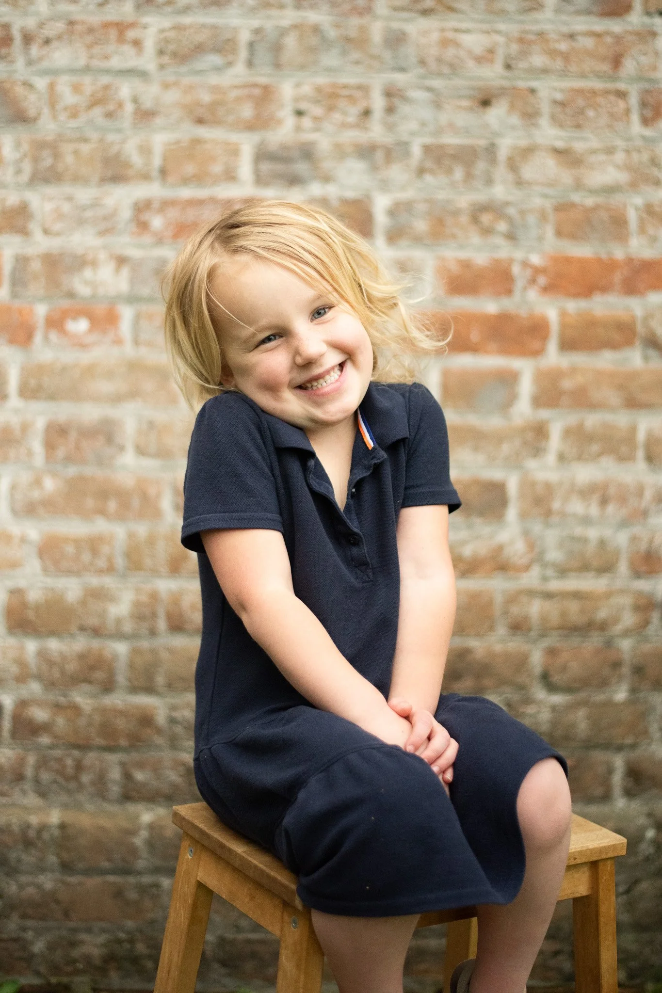 A young girl with blonde hair and blue eyes sitting on a wooden stool in front of a brick wall, smiling and wearing a navy polo shirt and navy shorts.