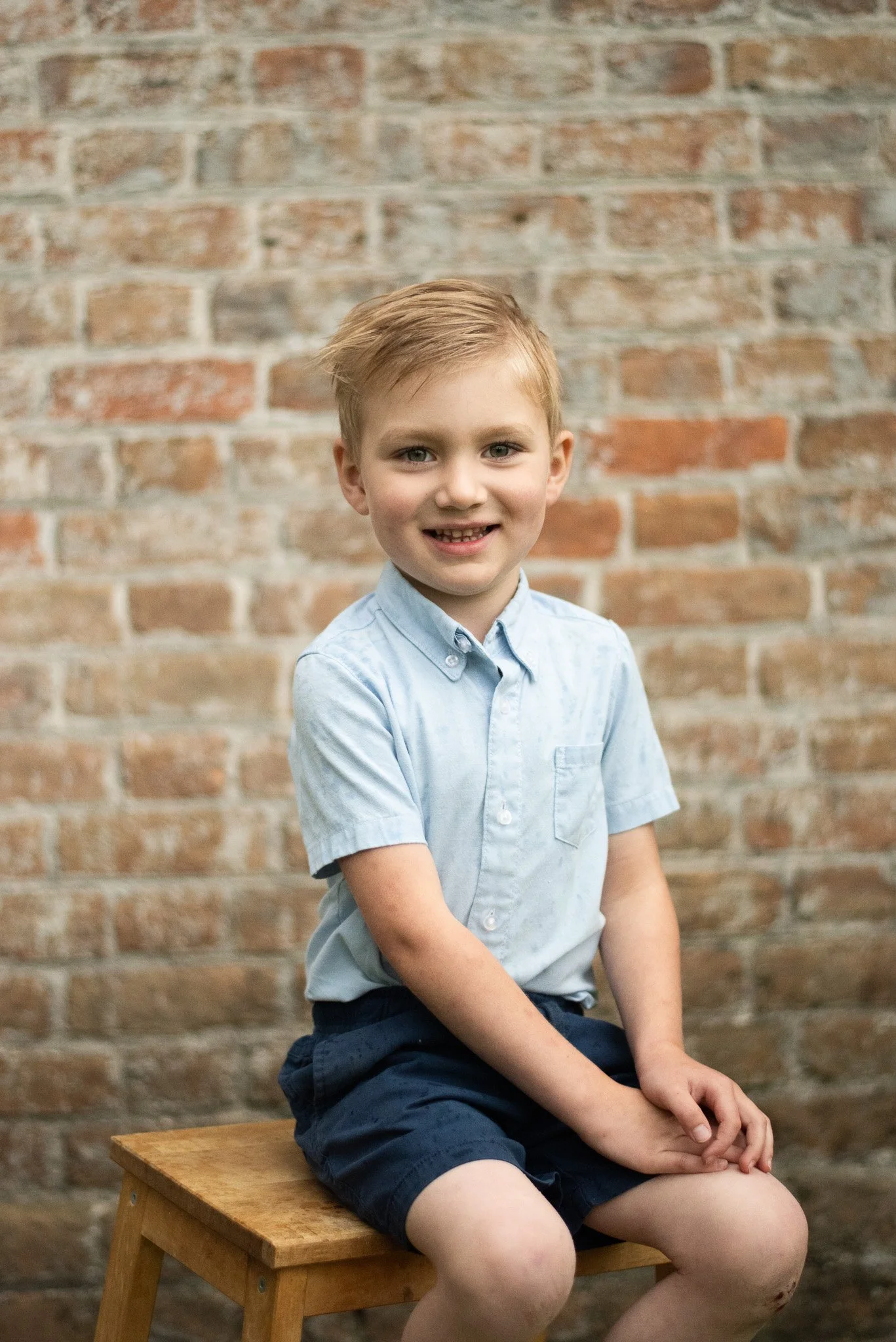 A young boy with blonde hair, blue eyes, and a big smile, sitting on a wooden stool against a brick wall background. He is wearing a light blue short-sleeved button-up shirt and navy shorts.