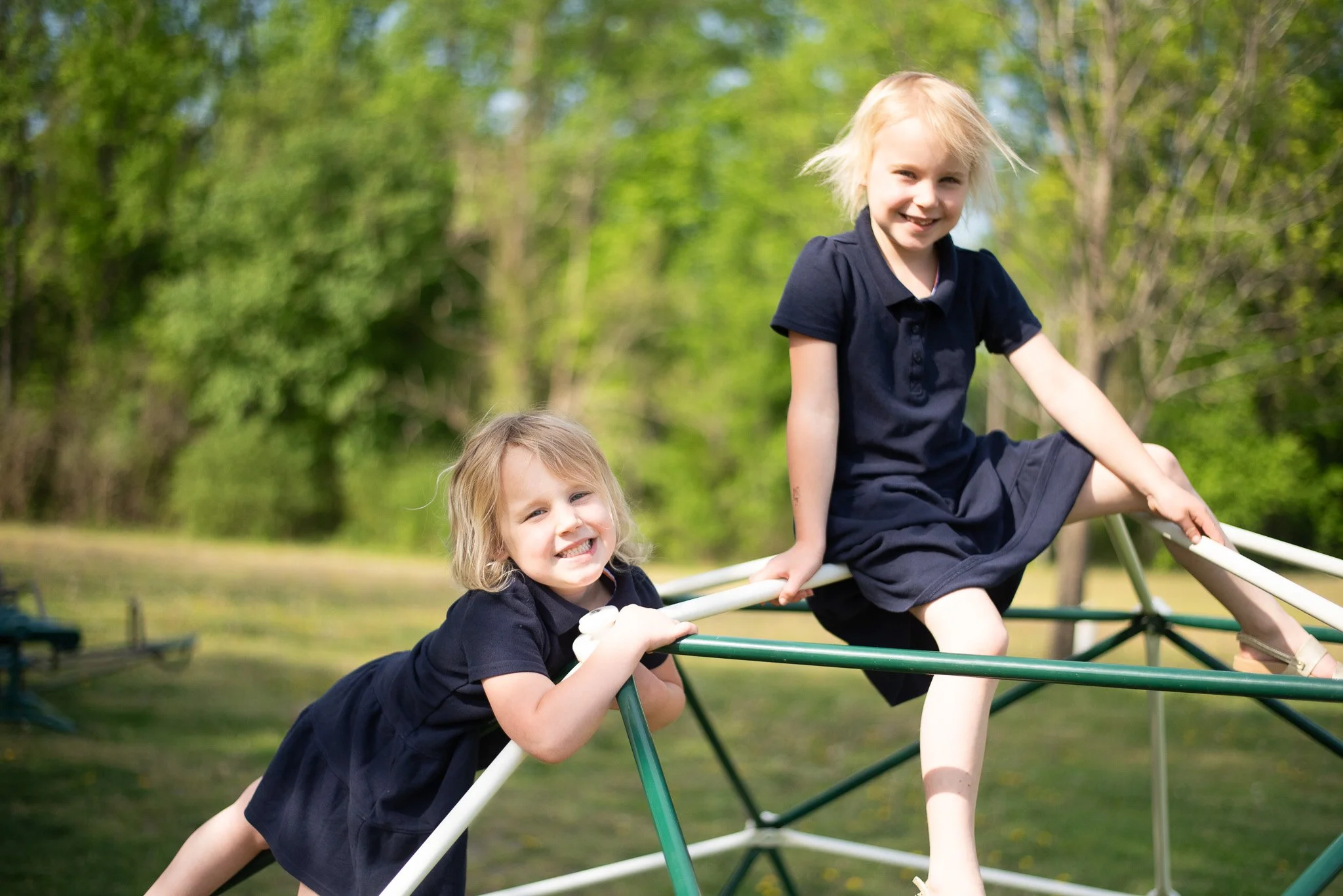 Two young girls with blonde hair playing on a jungle gym surrounded by green trees in the background.