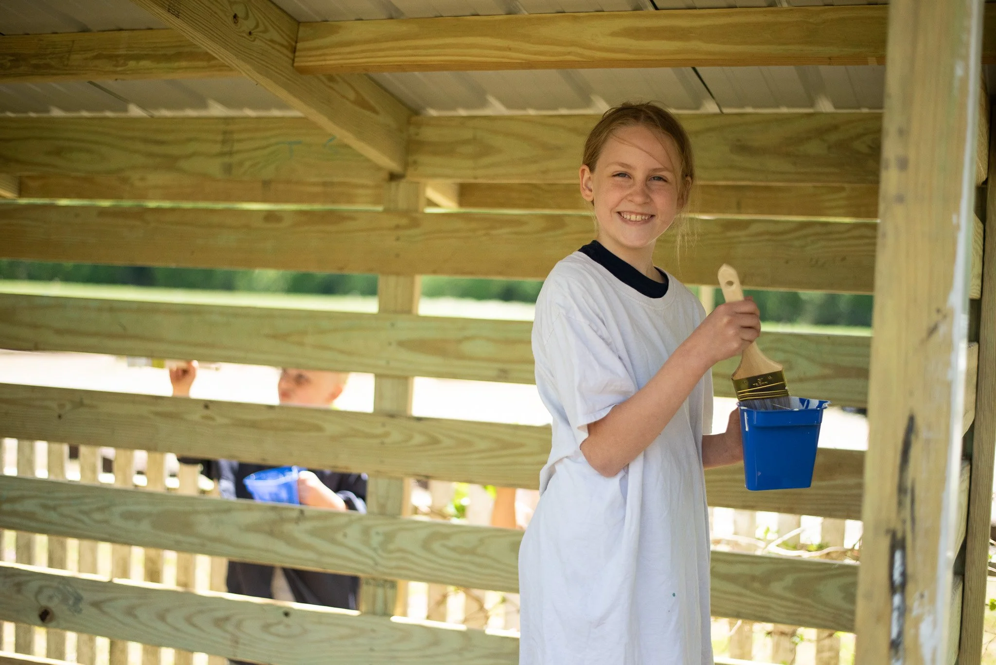 A young girl with red hair smiling and holding a paintbrush and a blue paint bucket, painting a wooden structure outdoors.