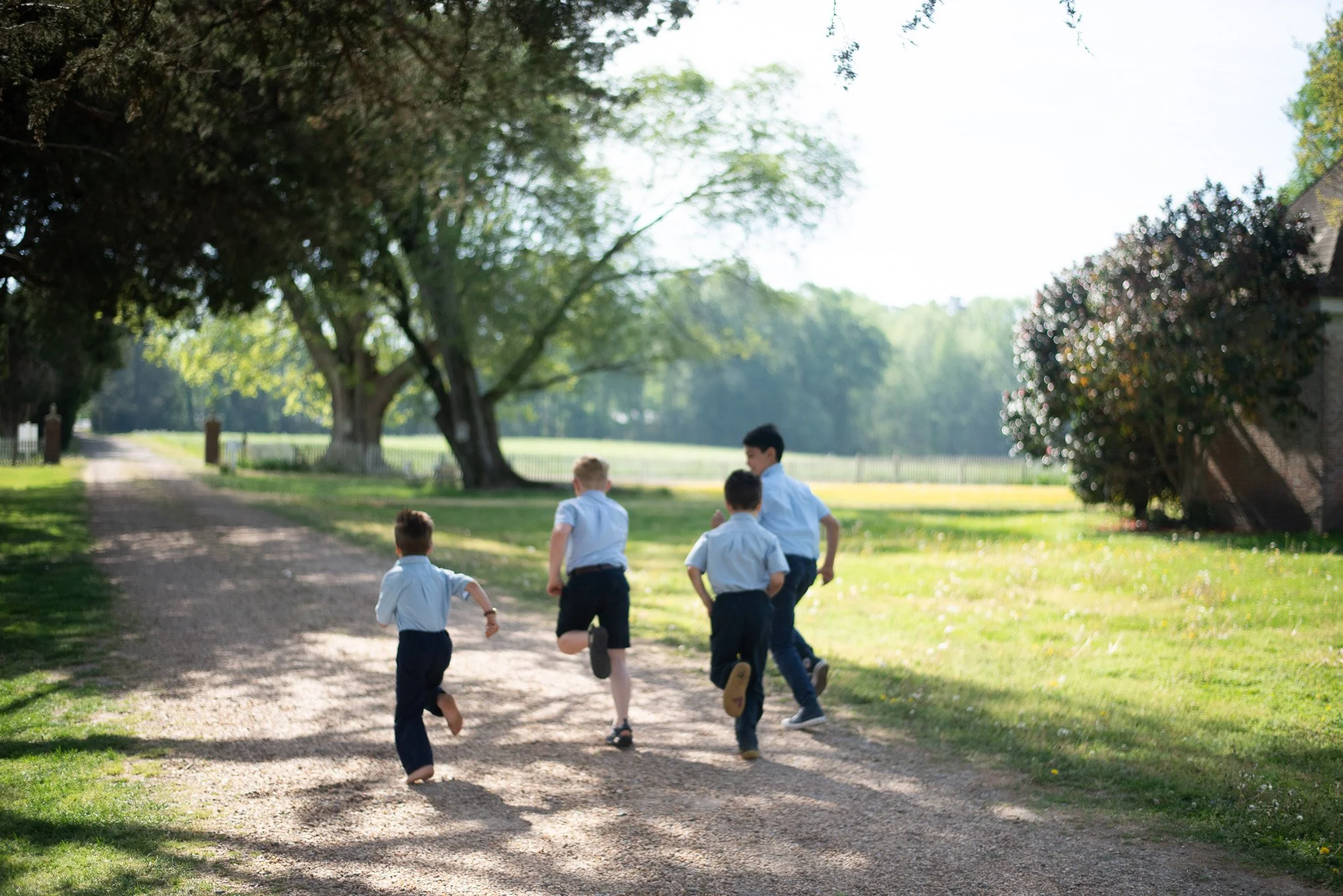 Group of children in school uniforms running on a dirt path in a park with large trees and green grass on a sunny day.