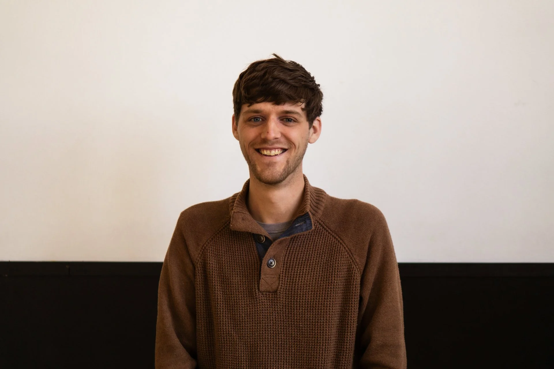 A young man with brown hair, blue eyes, and light skin smiling while standing in front of a plain white wall and a black panel at the bottom.
