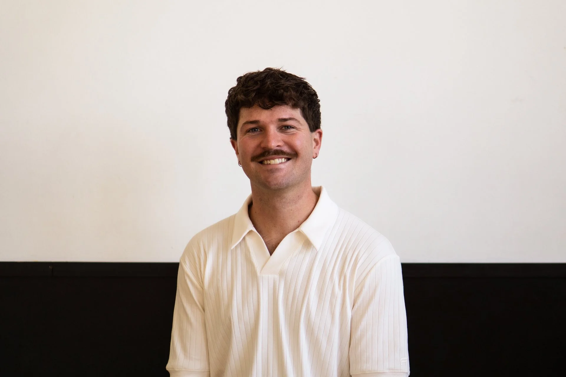 A smiling man with dark curly hair and a mustache, wearing a white collared shirt, standing in front of a white wall with a black lower panel.