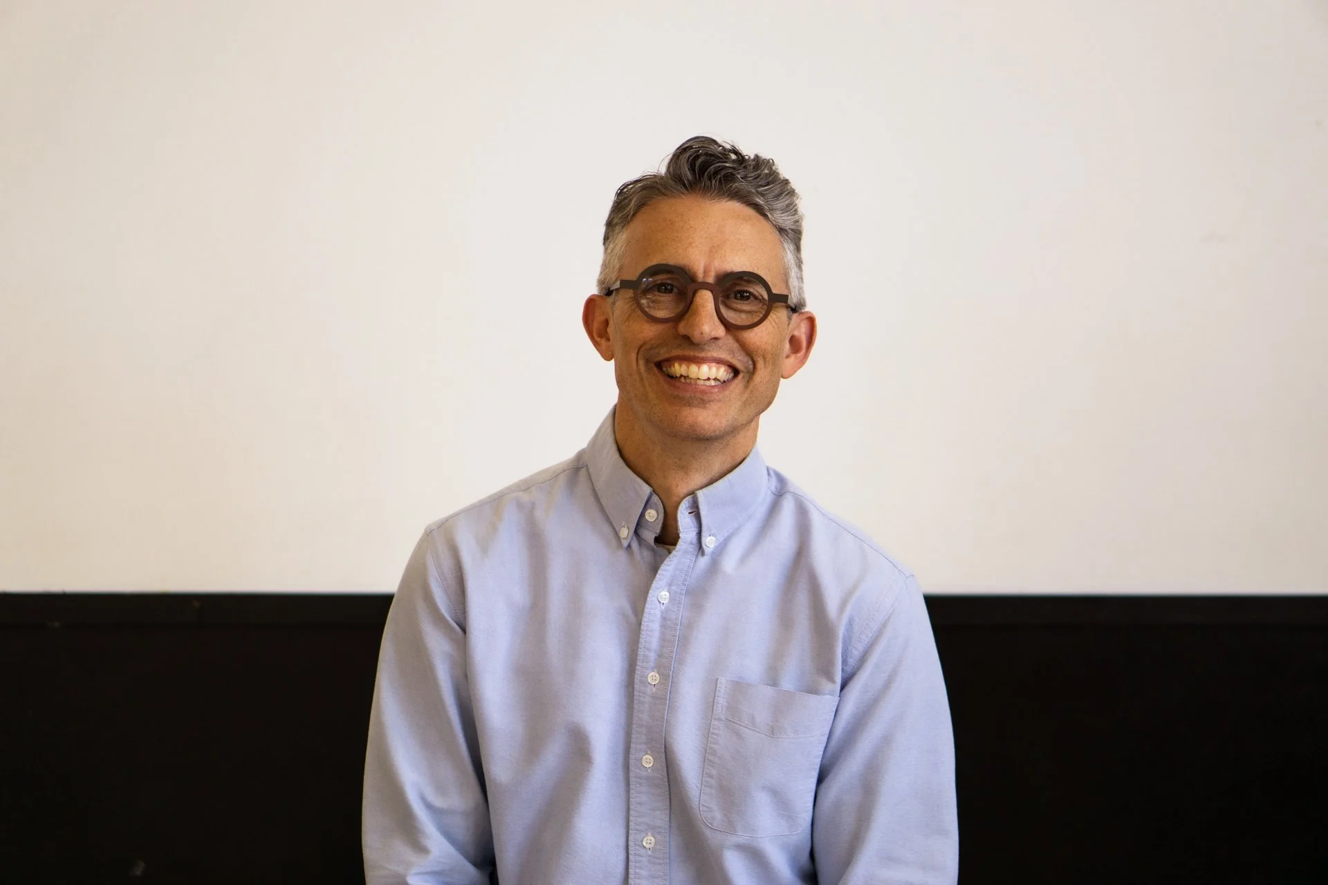 A man with gray hair, glasses, and a light blue button-up shirt, smiling and looking at the camera, sitting in front of a plain background.