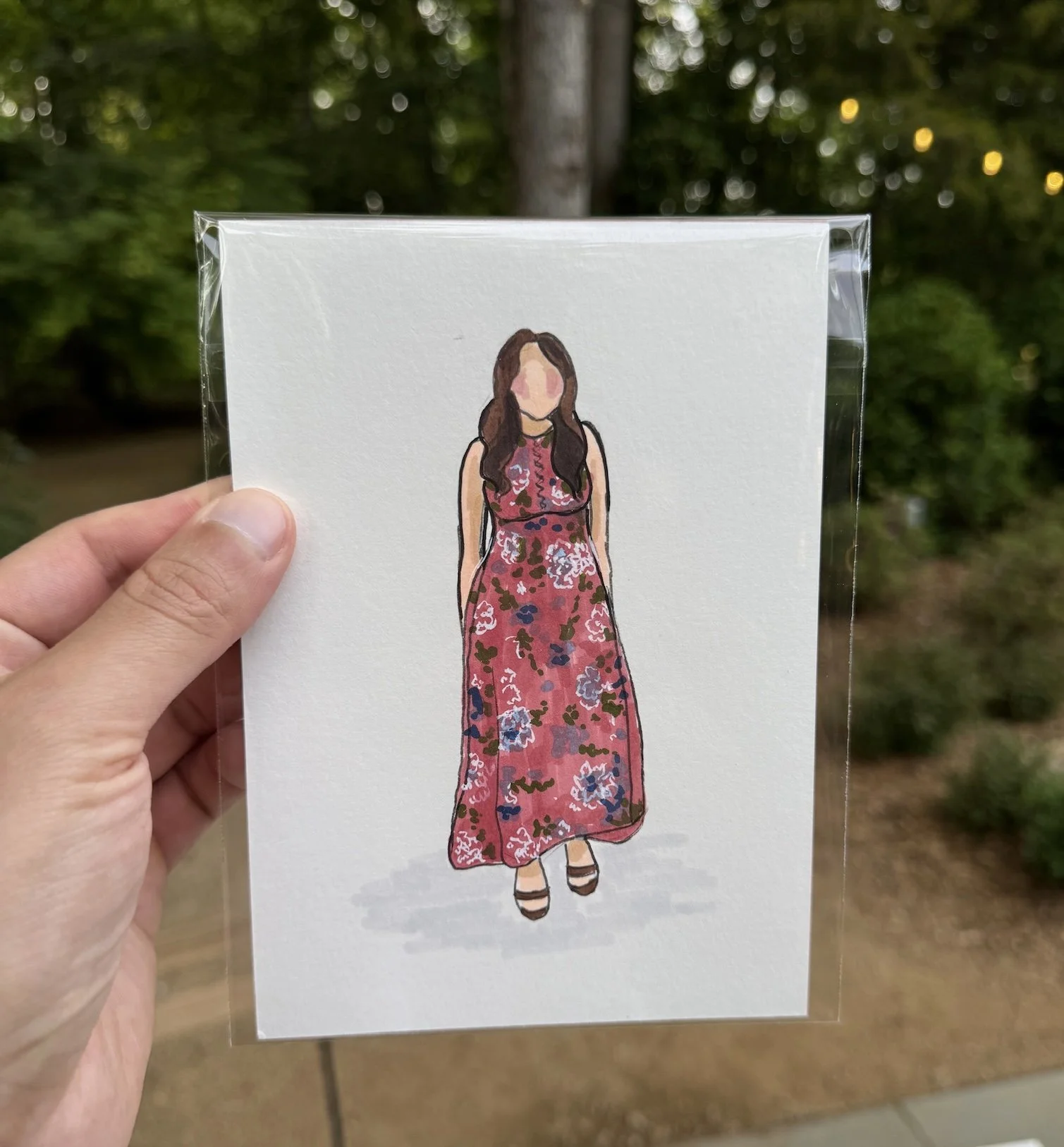 Hand holding a clear plastic sleeve with a watercolor illustration of a woman wearing a long, floral red dress, with a blurred outdoor background. Live guest portraits or wedding guest illustrations.