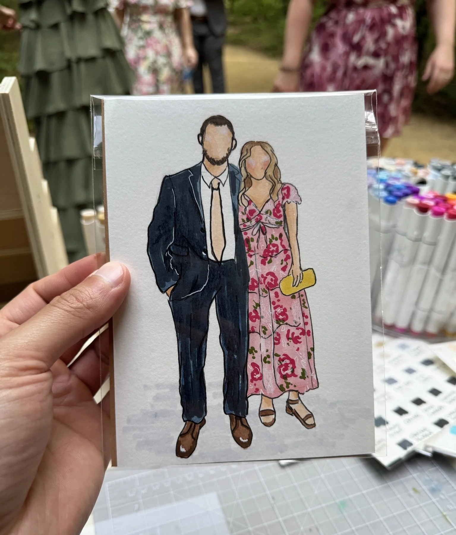 A person is holding an illustration of a man and woman standing together, the man in a suit and the woman in a floral dress, with no facial features. Live guest portraits and watercolor illustrations of guests.