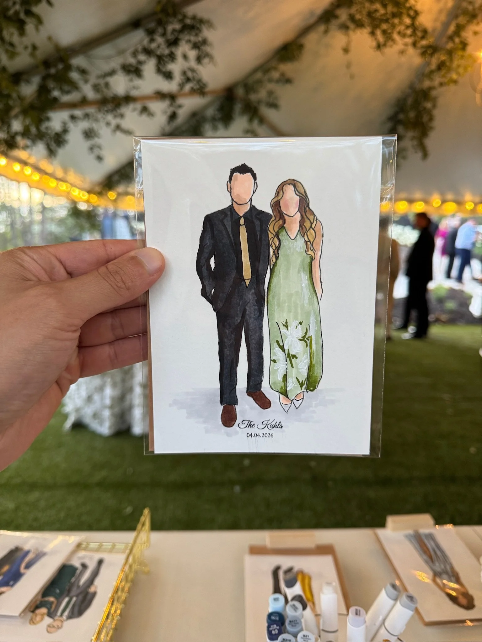 A wedding cocktail hour featuring a watercolor illustration of a couple in attendance, with the text "The Kuhls" and the date "04.04.2026" at the bottom. The drawing is held up in front of a decorated event space with string lights and greenery.