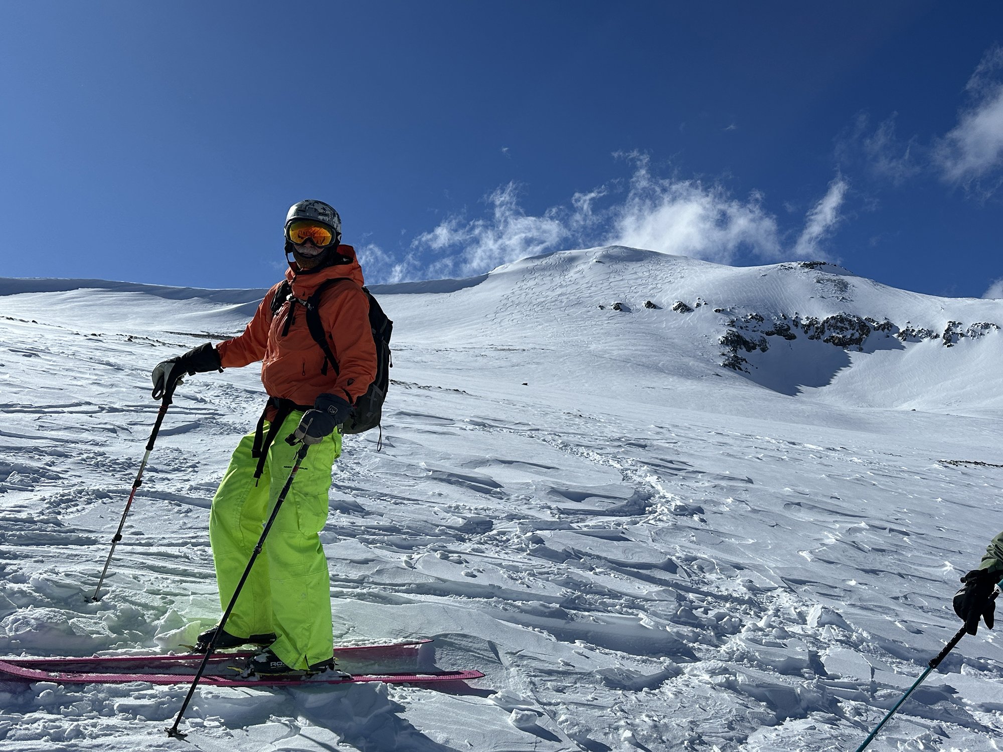 A person wearing bright green ski pants, an orange jacket, a helmet, and ski goggles stands on snow-covered terrain with ski poles, in front of snow-capped mountains and a blue sky.