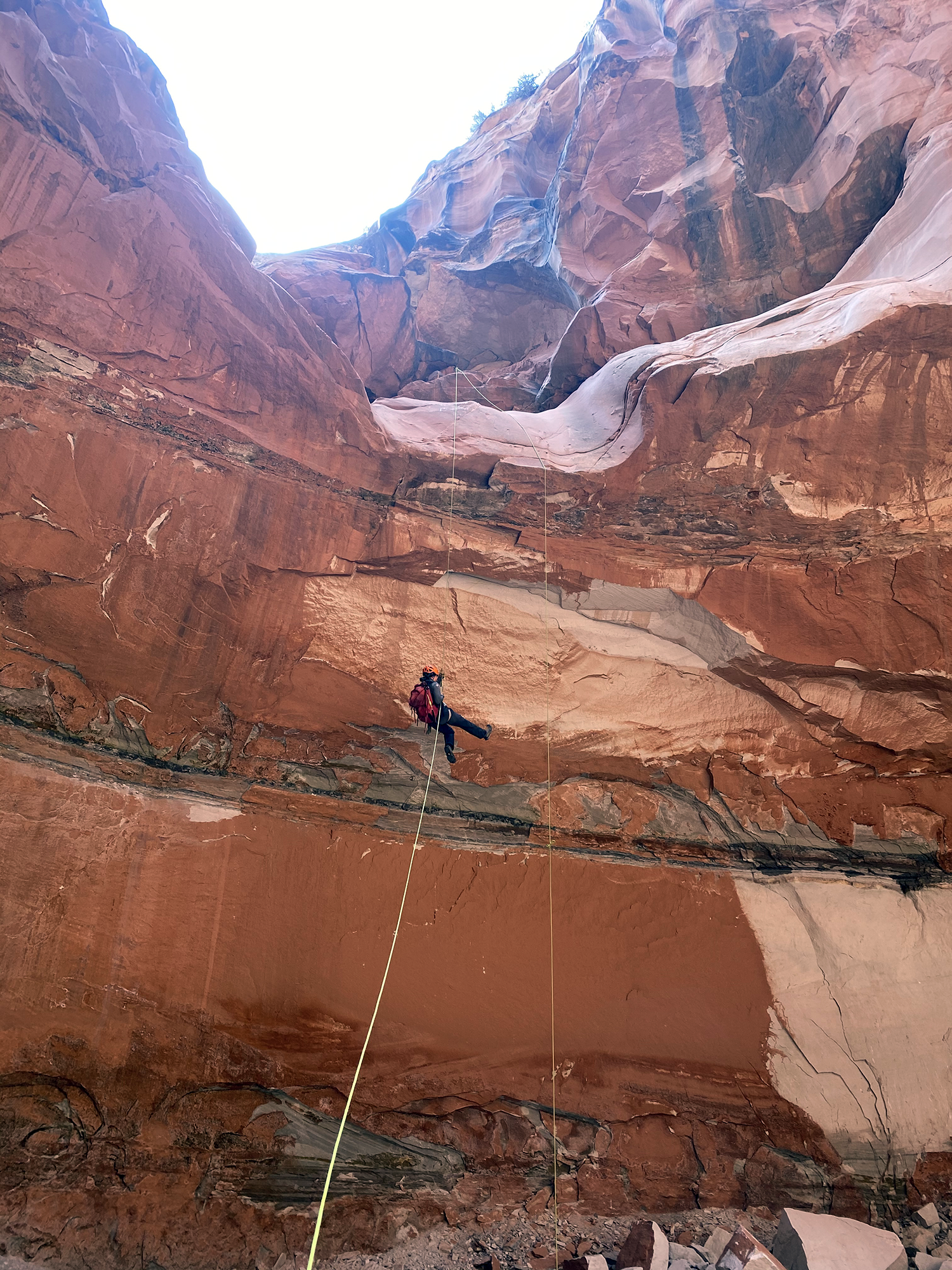 A person rappelling down a tall red rock canyon wall with a safety harness and rope in a desert landscape.