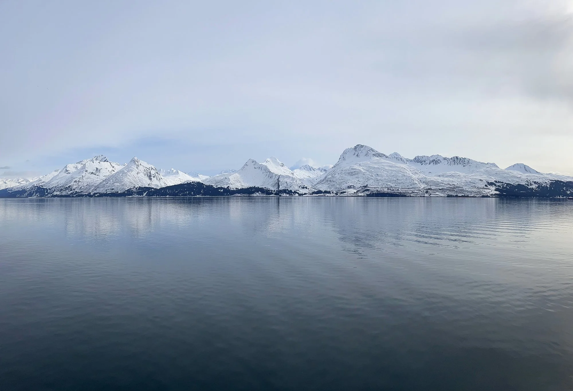 Snow-covered mountains reflected on calm water under a cloudy sky.