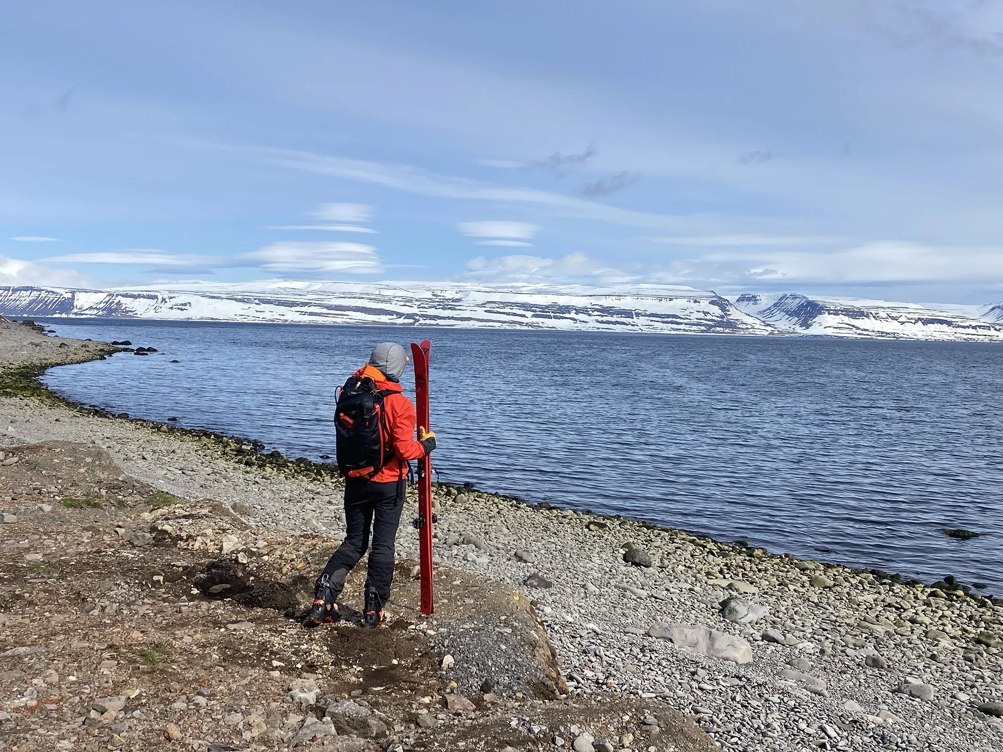 A person wearing a red jacket, black pants, and ski boots carrying red skis over their shoulder, walking along rocky shoreline by a body of water with snow-covered mountains and cloudy sky in the background.