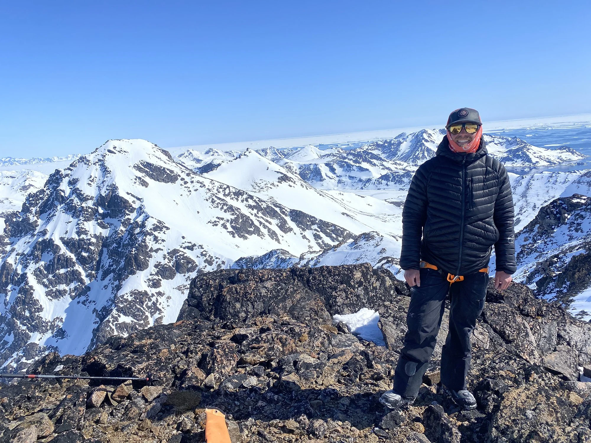 A person dressed in black outdoor gear, including a puffy jacket, pants, and hiking shoes, stands on rocky terrain at a snowy mountain summit. They are wearing a helmet, reflective sunglasses, and a neck gaiter, smiling with snow-capped mountains in the background under a clear blue sky.