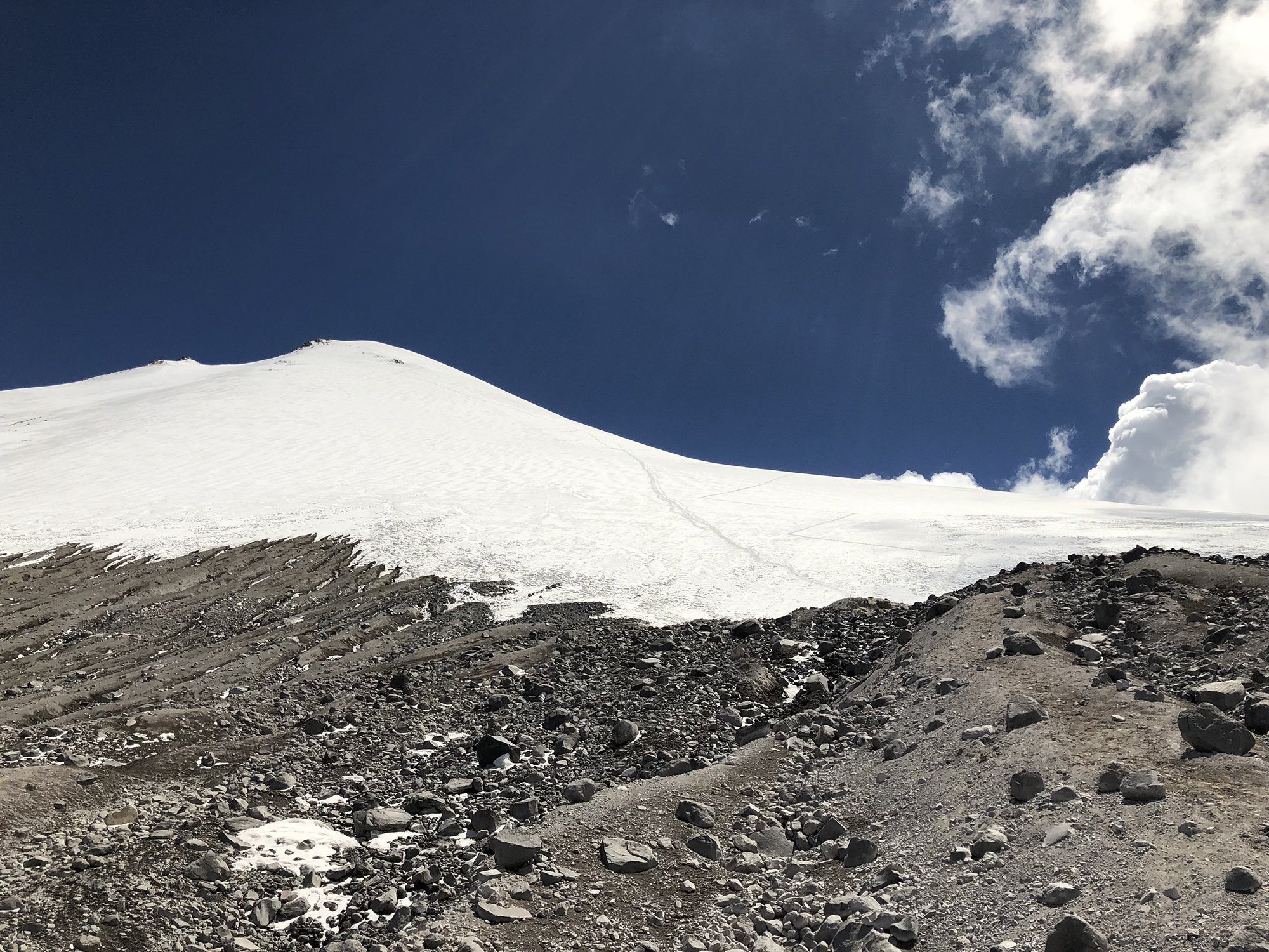 Snow-covered mountain slope with a rocky terrain at the base, under a blue sky with scattered clouds.