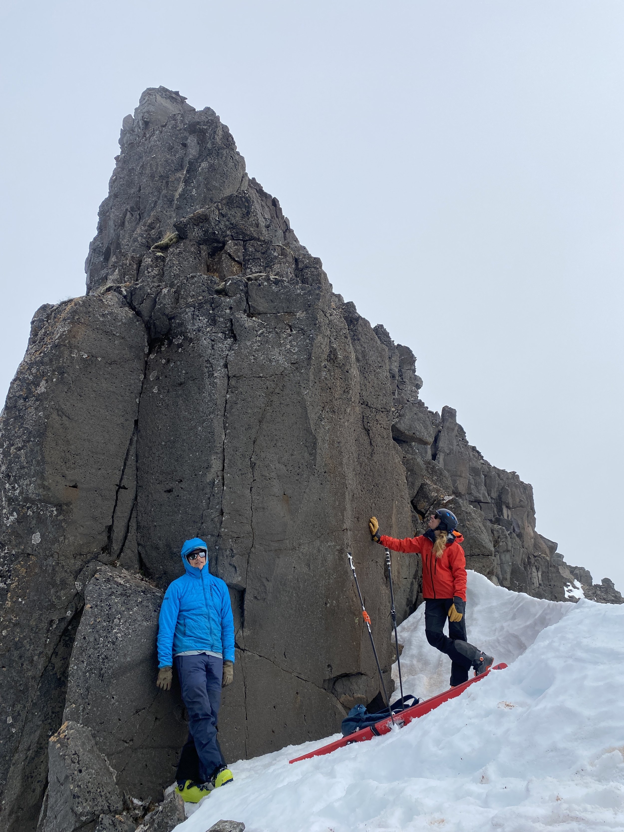 Two climbers in bright outdoor gear standing next to a large rocky cliff on snow-covered ground, one is holding a climbing pole and the other is leaning on the rock with a helmet, bikes, and snow gear nearby.