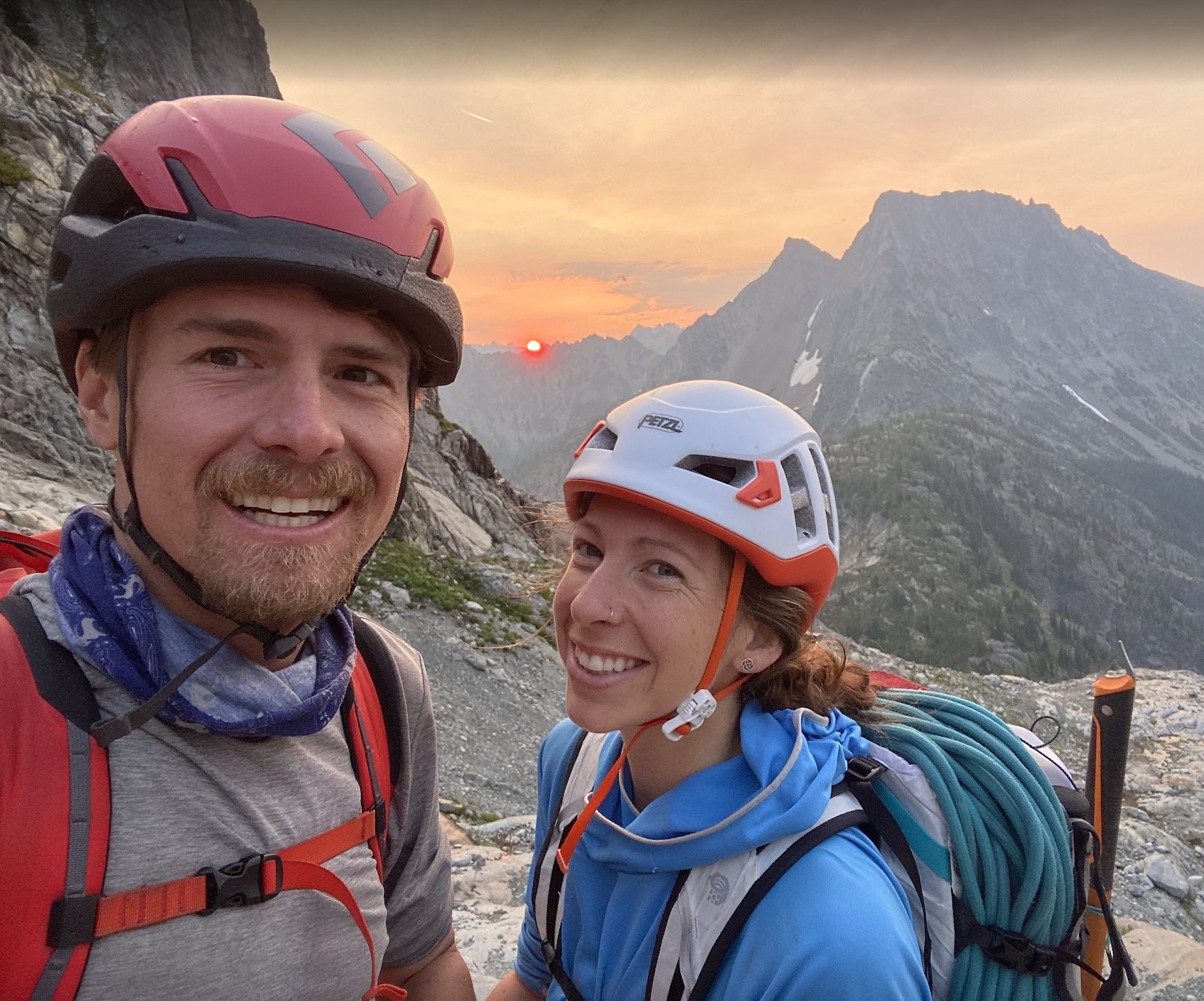 Two hikers taking a selfie in a mountainous landscape during sunset, wearing helmets and backpacks.