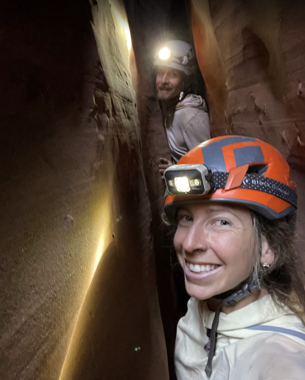 Two women in helmets with headlamps exploring a narrow, underground slot canyon with red rock walls, smiling at the camera.