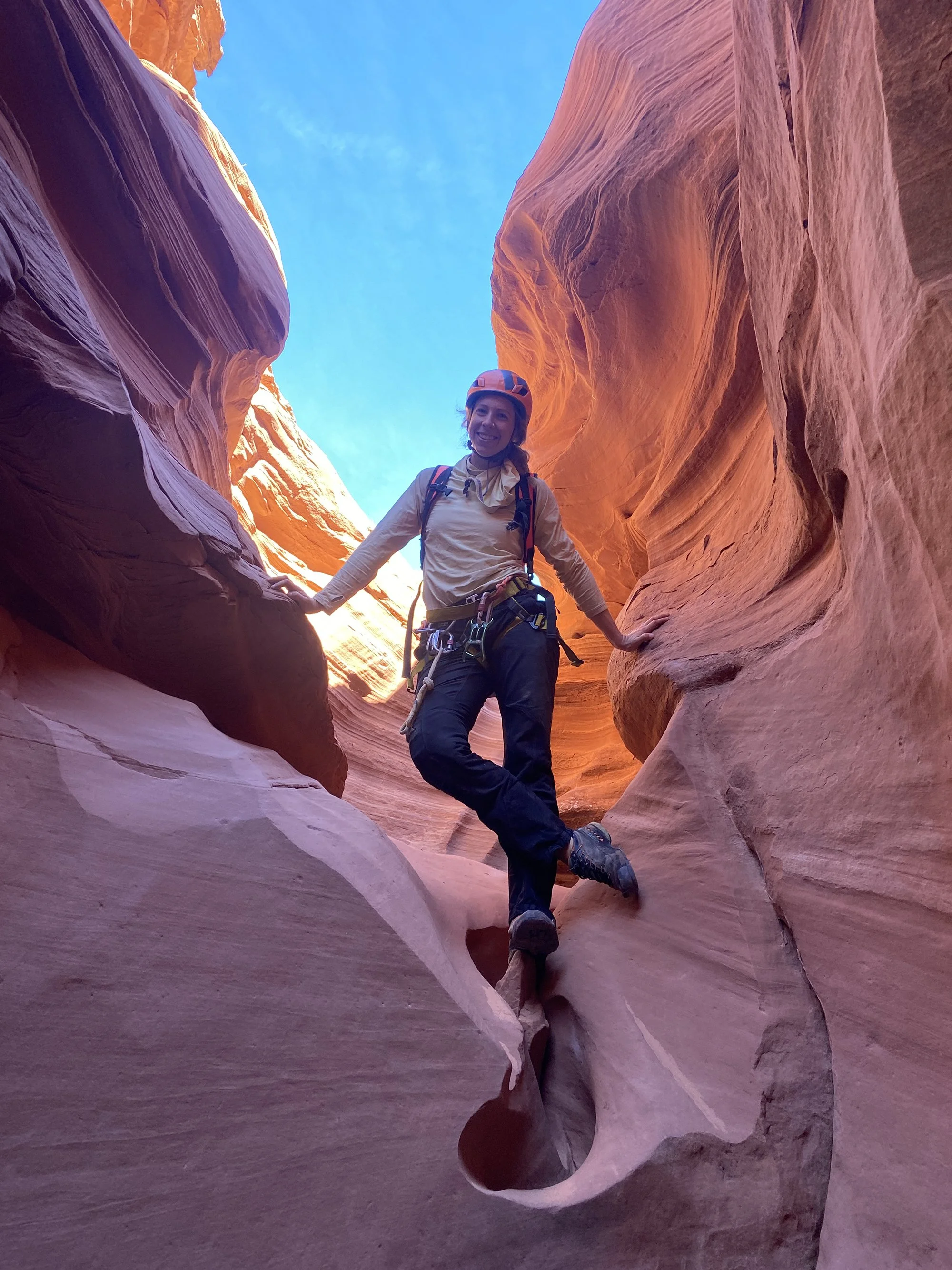 A woman in outdoor climbing gear, including a helmet, harness, and backpack, stands among red canyon walls with a bright blue sky above, smiling at the camera.